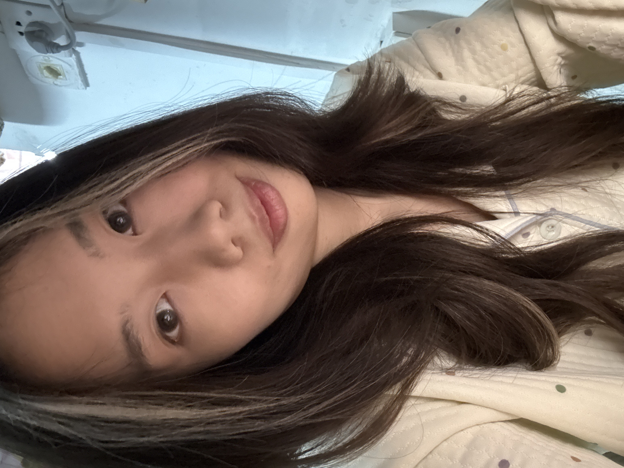 A close-up portrait shows a young East Asian female positioned in an indoor setting within Nan'an District, China. The subject, with long, dark brown hair featuring lighter streaks, faces forward, looking directly at the camera with a subtle, closed-mouth smile. She wears a quilted, light-colored shirt or pajama top adorned with small, multi-colored polka dots and light blue piping along the collar and button placket. The background, visible primarily behind her left side, reveals a light blue or white wall with a white electrical wall outlet and a gray plug inserted into one of its receptacles.