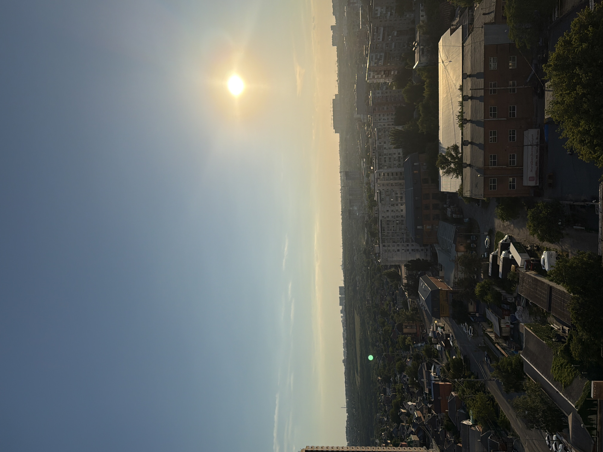 The image is a vertical, high-angle shot capturing an urban landscape at sunset. The sun, a bright, yellowish orb, is prominently featured in the upper left quadrant of the sky, casting a warm glow and creating lens flare. The sky transitions from a clear blue at the top to a hazy yellow closer to the horizon, suggesting atmospheric conditions typical of sunset.
The scene depicts a sprawling city with numerous buildings of varying heights, including apartment blocks and what appear to be industrial or commercial structures. In the foreground, on the right side of the image, a large brick building with a light-colored roof is visible, its windows reflecting the sunlight. Trees, some with lush green foliage, are scattered throughout the urban environment. A road with parked vehicles is also visible, indicating a lived-in area.
Further into the distance, more buildings and a strip of green, possibly a park or undeveloped land, can be seen before the horizon is met by a hazy, muted cityscape. The overall impression is one of an active urban area bathed in the soft, warm light of the setting sun. There is no discernible activity or people in the image. A piece of text is visible on a white sign attached to the side of the brick building, but it is too blurry to be read. The location is identified as Ovoshchnoy, Russia.