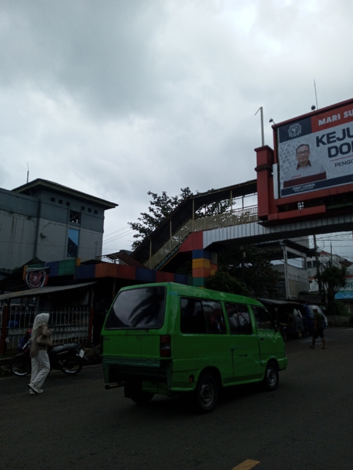 The image captures a bustling street scene in an urban or semi-urban area, likely in Indonesia, under a heavily overcast sky. The lighting is diffused and grey, suggesting either an early morning, late afternoon, or simply a very cloudy day.
In the foreground, a dark asphalt road occupies the bottom portion of the frame. A bright green microbus or minivan, possibly an older model, is the most prominent vehicle, positioned slightly to the right of the center, with its rear facing the viewer. Its windows reflect the cloudy sky. To the left, a person wearing a light-colored hijab, a light brown top, and white pants with white sneakers is walking from left to right along the side of the road. Two motorcycles are parked near a fence behind this person. Further back on the right side of the road, several other people are visible, appearing somewhat blurry due to movement, with one individual wearing a backpack.
The midground features a complex arrangement of buildings and structures. On the left, a multi-story building with a grey and light grey facade and square windows stands. Below it, a smaller, low-roofed structure, possibly a shop or stall, adds a splash of color with its base painted in vibrant blocks of red, blue, yellow, and green, topped with a dark corrugated roof and fronted by a metal fence. This colorful structure appears to form the base of a pedestrian overpass.
The pedestrian overpass itself is a prominent feature, extending from the left towards the right, spanning over the road. It has a yellow railing and black support beams along its stairs and walkway, consistent with the colorful base structure. Behind the overpass and buildings, some green foliage from trees is visible, offering a touch of nature amidst the urban setting.
In the background, on the right side of the frame, a large red and white billboard dominates the upper portion. It features a portrait of a man wearing glasses and a suit, who appears to be a public figure or politician. Visible text on the billboard includes:
- "MARI SU..." (likely part of "MARI SUKSES" or a similar phrase, meaning "Let's succeed").
- A logo with some text below "MARI SU...".
- Large bold text: "KEJU..." and below it "DO...".
- Below the portrait, smaller text names are visible: "HARY TANOESUDIBJO" and "RIDOY SAHRUL".
- Partially obscured large text below the names: "PENGGO..." (possibly "PENGGOLONGAN" or related).
The scene depicts everyday life with pedestrians and vehicles moving under an overcast sky, suggesting a typical day in this bustling area. No strong emotions are discernible, and the overall atmosphere is one of routine activity.