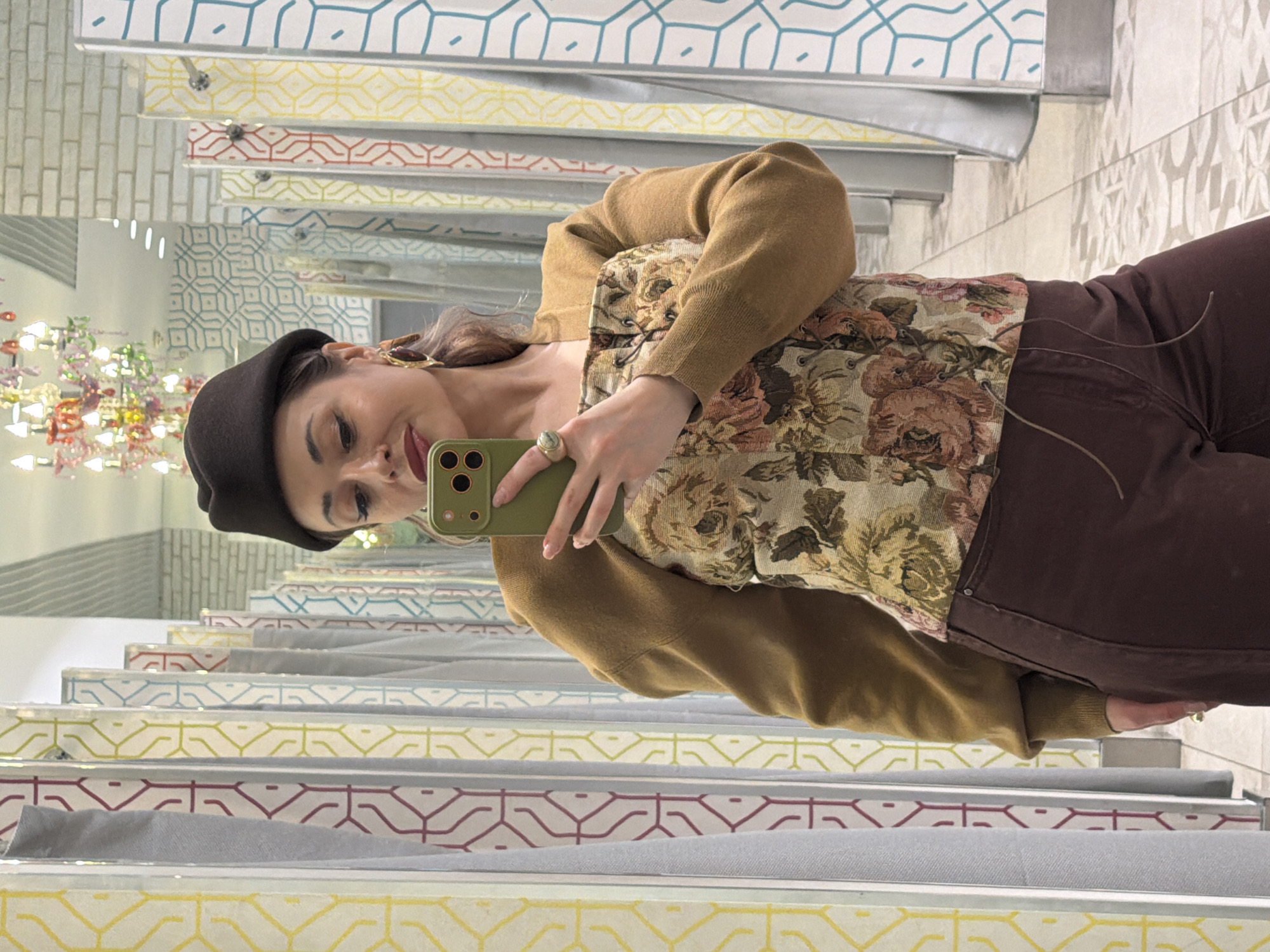 A woman is taking a selfie in front of a mirror. She is wearing a beret, floral top with a mustard-colored cardigan, and brown pants. She is holding a green smartphone in her left hand and has a ring on her left ring finger. The mirror shows reflections of clothing stalls with patterned partitions. In the top left of the frame, a colorful chandelier can be seen in a mirror. The location appears to be a dressing room in Krasnoyarsk, Russia, due to the reflections of store fitting room interiors.