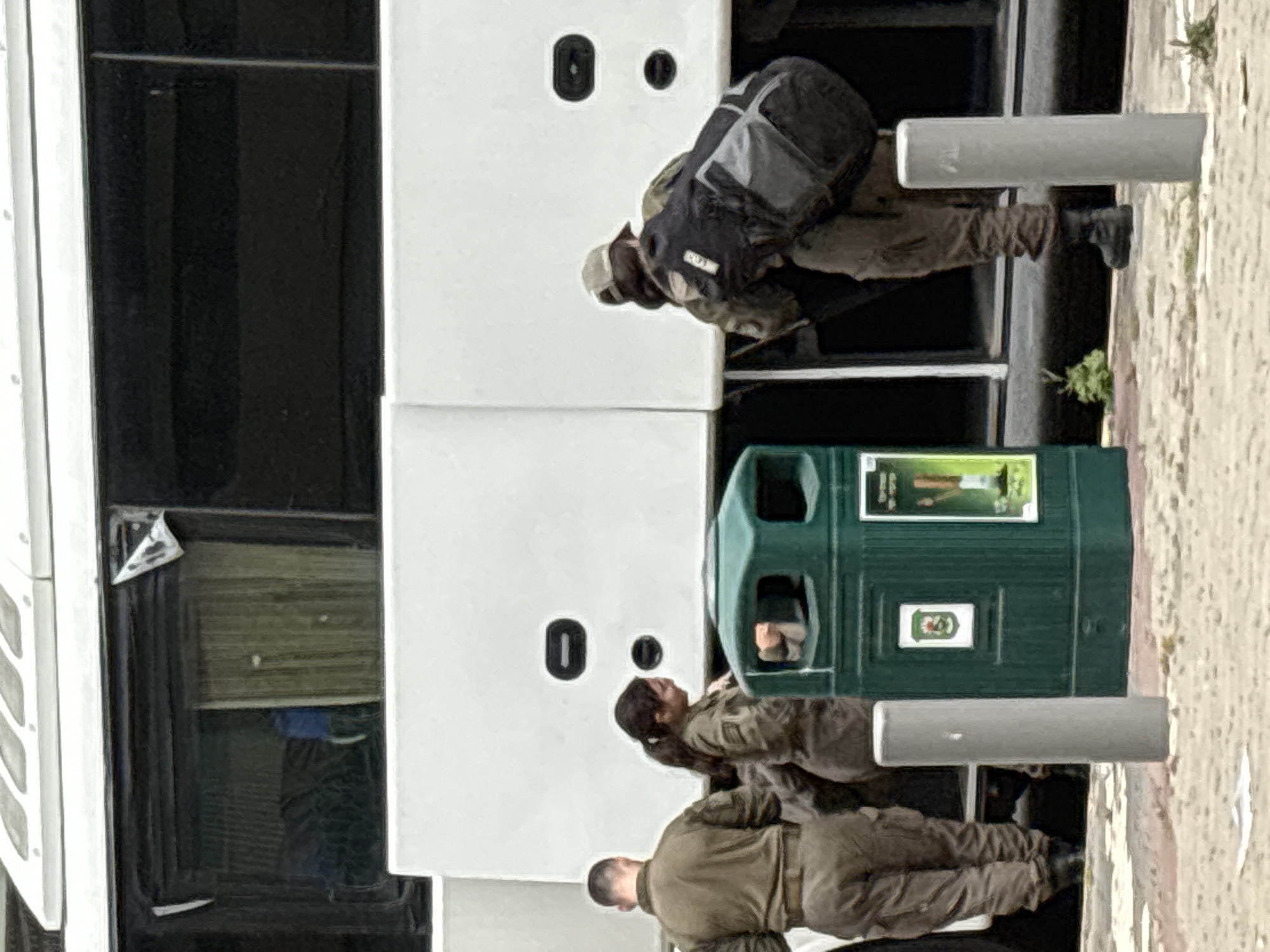 This image, taken in Ramat Gan, Israel, captures an outdoor scene featuring four individuals, a white bus, and a dark green public waste bin. The photograph appears to be oriented vertically, suggesting it was originally taken with the camera rotated 90 degrees clockwise.
Three individuals are clearly visible in the foreground, dressed in olive green or tan uniforms, characteristic of military or security personnel. One person, seen from behind, is wearing a large dark backpack and is positioned near the side of the white bus, possibly in the process of loading or unloading items. A woman with dark hair tied back stands near a dark green public waste bin, facing towards the bus. Another man, also in uniform, is seen from behind in the lower part of the frame. A fourth person is faintly visible through one of the bus's dark tinted windows.
The large white vehicle is a bus, featuring dark windows and two black, oval-shaped fixtures on its side. Adjacent to the bus stands the dark green waste bin, which displays two stickers: a smaller white square with a green recycling symbol and a larger image above it that appears to show a person in a green field, possibly an advertisement or public awareness message. The ground is paved, indicating an urban or semi-urban setting, potentially a bus stop or public area. Two grey cylindrical bollards or posts are also present in the scene. The overall lighting suggests it is daytime, possibly under overcast conditions.