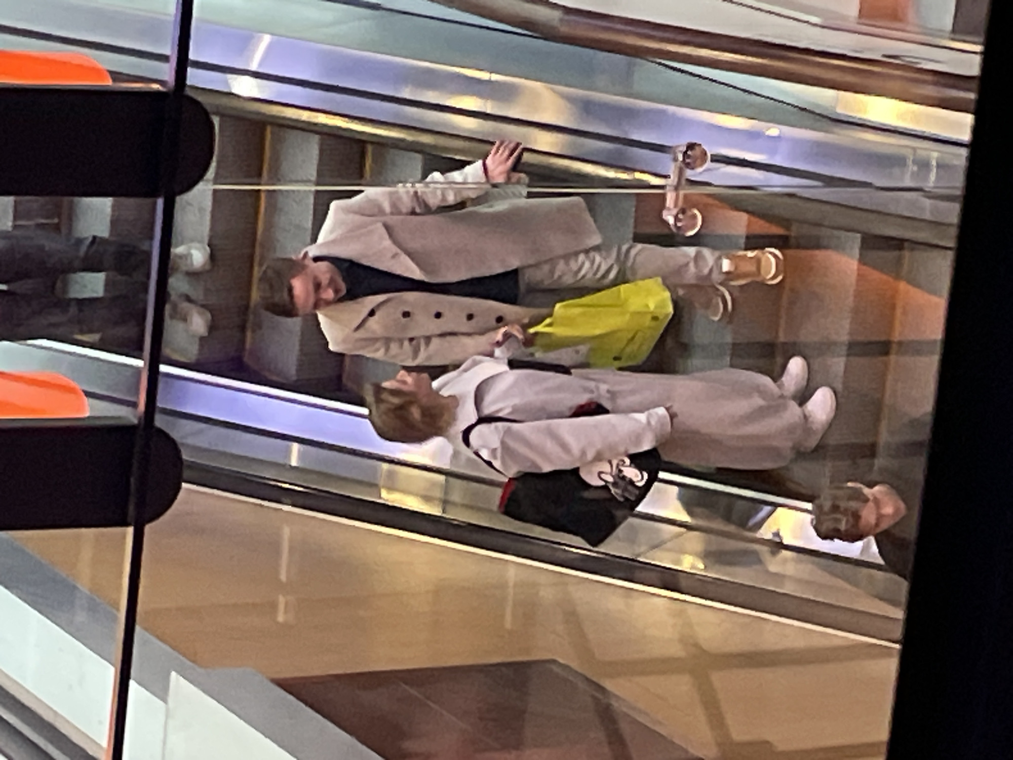 The image shows two individuals, a male and a female, on an escalator in what appears to be a shopping mall or public transport hub. The male is wearing a light-colored trench coat, dark shirt, and light pants with brown shoes. He is holding a yellow bag. The female is wearing a light-colored overcoat and pants with white shoes, holding a black bag. The escalator is located in a bright, modern indoor space, potentially in Saint Petersburg, Russia. The reflection on the glass shows another person below them.