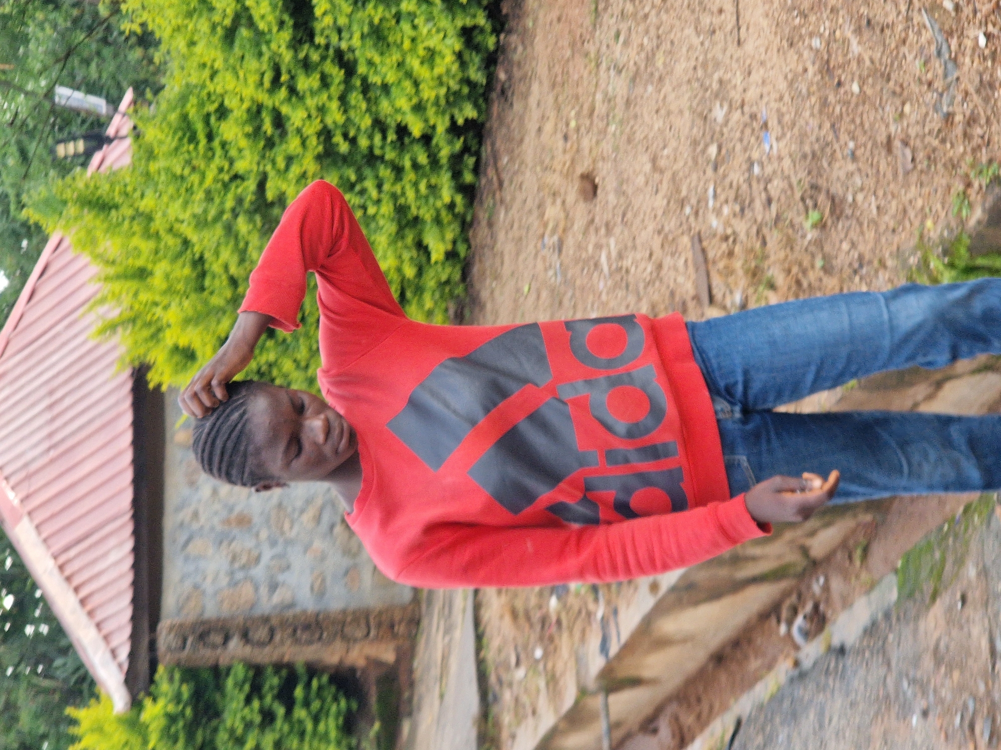 A young person with braided hair stands in front of a lush green hedge and a building with a red, sloped roof. The person is wearing a red long-sleeved sweatshirt with a large black Adidas logo on the front, and blue denim jeans. Their right hand is raised to their head, and their gaze is directed downwards. The ground is covered with dirt and scattered debris. The building appears to be made of stone and has a somewhat weathered appearance. This scene is set in Kabong, Nigeria.