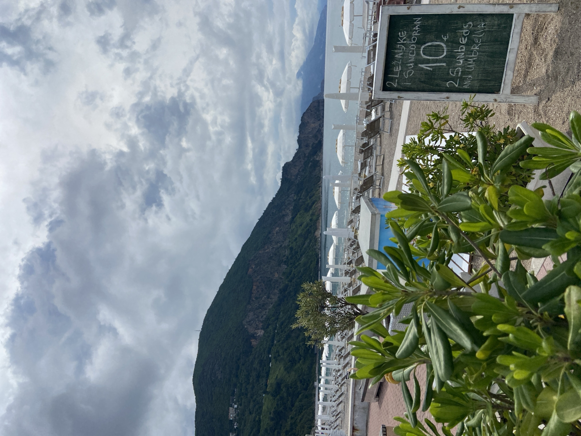 The image shows a coastal scene in Prijevor, Montenegro. In the foreground, a sign advertises "2 lezaljke i suncobran 10€" which translates to "2 sunbeds and umbrella 10€". The sign is positioned next to lush green foliage, suggesting a resort or beachfront property. Beyond the foliage and sign, rows of white sunbeds with umbrellas line the shore. A strip of water and then a large, forested mountain are in the middle ground. The sky above is filled with an array of cloudy formations.