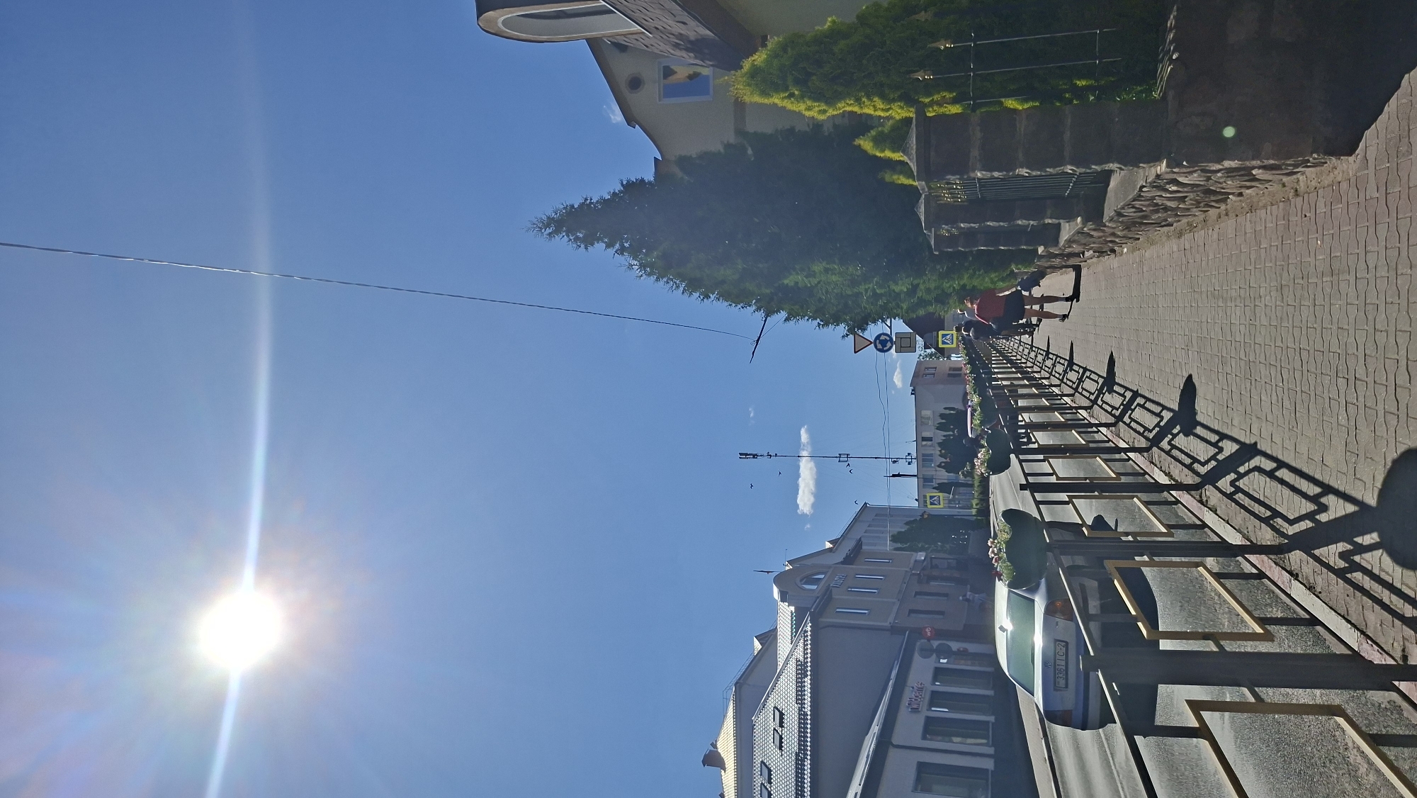 The image captures a sunny day on a city street in Pastavy, Belarus. A bright sun with a lens flare is prominent in the upper left corner of the clear blue sky. A dark power line stretches diagonally across the sky. The scene depicts a sidewalk paved with interlocking bricks, lined by a metal fence with yellow accents. On the sidewalk, two individuals are walking away from the camera. The person in front is wearing a red top and dark shorts, while the person behind them is wearing a dark top and shorts. Several decorative evergreen trees and stone pillars flank the sidewalk. Across the street, there are buildings with tiled roofs, some appearing to be commercial establishments. A light-colored car with a license plate is driving on the asphalt road, which is separated from the sidewalk by the fence. Planters with flowers are visible along the fence. Traffic signs are present on the side of the road, including pedestrian crossing signs and a circular sign with a blue background and a white arrow. The overall atmosphere suggests a normal day in a European city.
