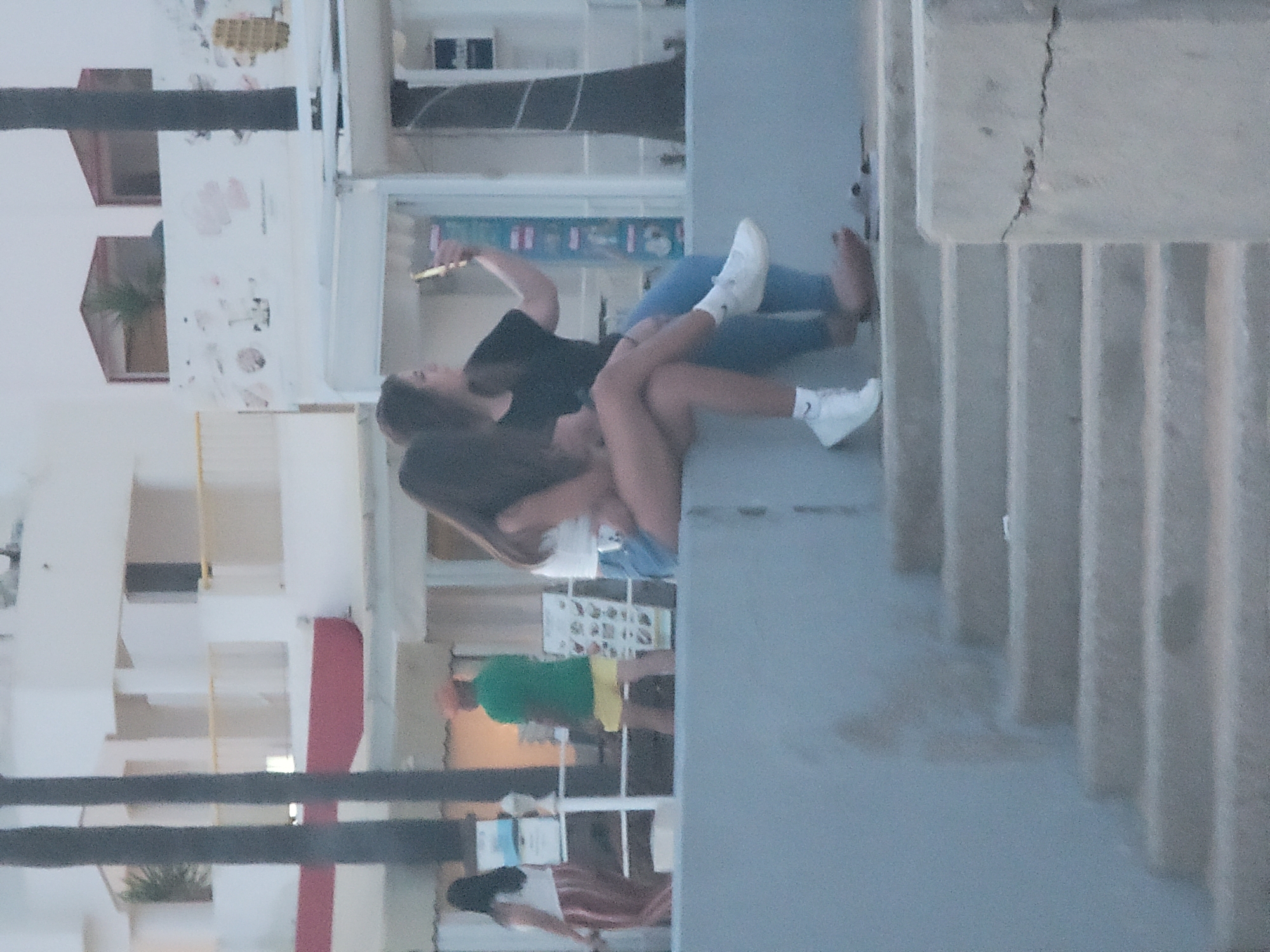 Two young women are seated on a concrete ledge overlooking a public walkway. The woman on the right is wearing a black t-shirt and blue jeans, and is holding up a smartphone, seemingly taking a selfie. The woman on the left is wearing a white crop top and denim shorts, with white sneakers. The background shows a building with balconies, palm trees, and a street scene with people and a food stall. The overall lighting suggests it is late afternoon or early evening.