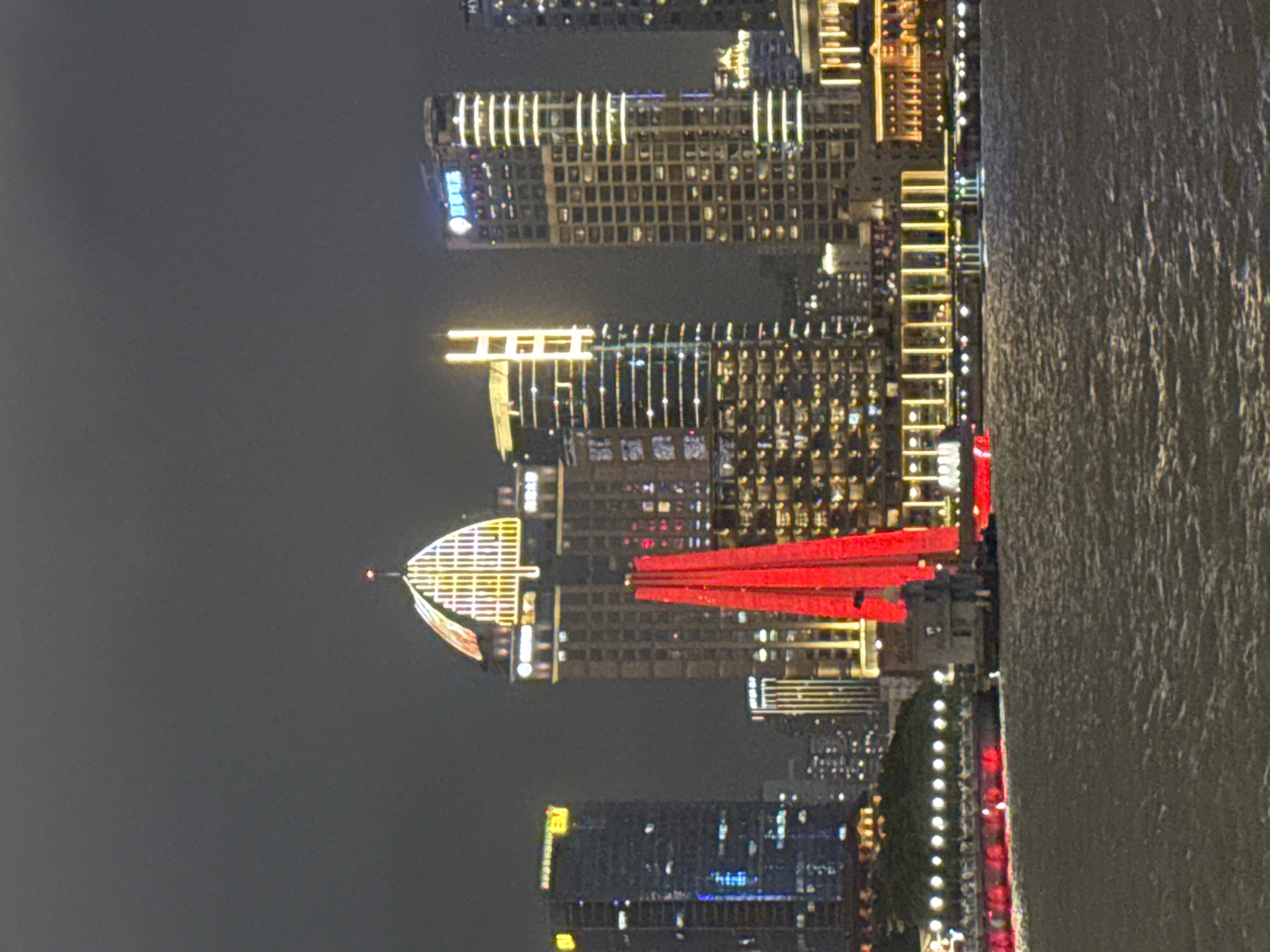 The image shows a nighttime view of the Pudong skyline in China, with several modern skyscrapers illuminated against a dark sky. The foreground features a body of water, likely a river, with ripples on its surface. In the middle ground, a distinct red, triangular structure, possibly a piece of public art or architectural element, stands prominently. Behind it, buildings are lit with various patterns of lights, including vertical strips and grid-like facades. Some buildings display illuminated signage, one in blue characters. The overall impression is of a vibrant and illuminated urban waterfront at night.