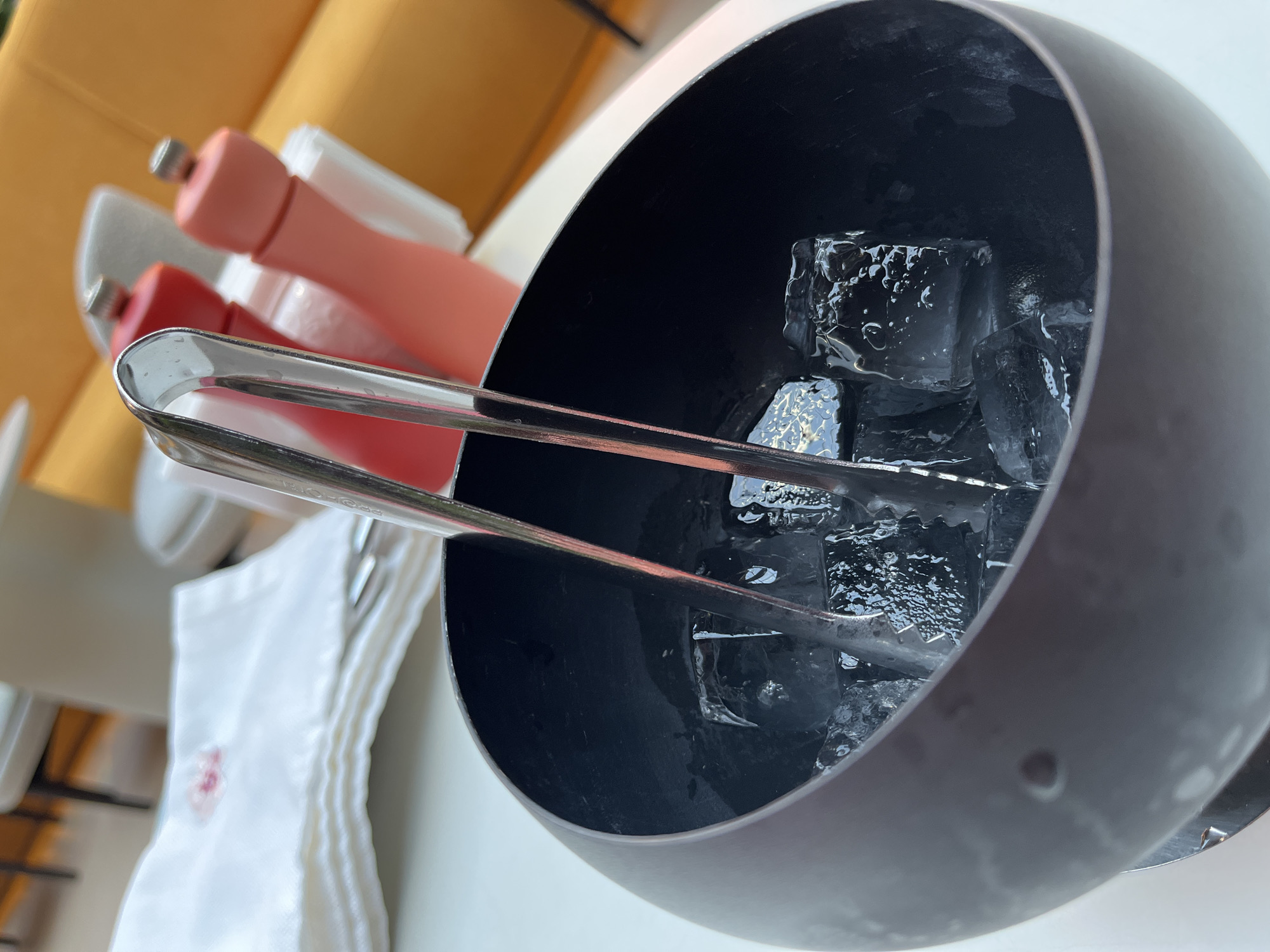 A close-up, eye-level shot focuses on an ice bucket filled with ice cubes and a pair of metal tongs. The ice bucket is dark, wide, and appears to be made of metal or a similar material. The tongs are silver and are positioned in the center of the ice bucket, with their tips submerged in the ice.
In the background, out of focus, are a stack of white napkins, one of which has a faint pink stain. Two salt and pepper grinders, one red and one coral pink, stand behind the napkins. Further in the background, a portion of a bright orange sofa is visible, along with abstract shapes of furniture and architectural elements suggesting an indoor dining or lounge setting. The overall lighting is bright, possibly from natural light.