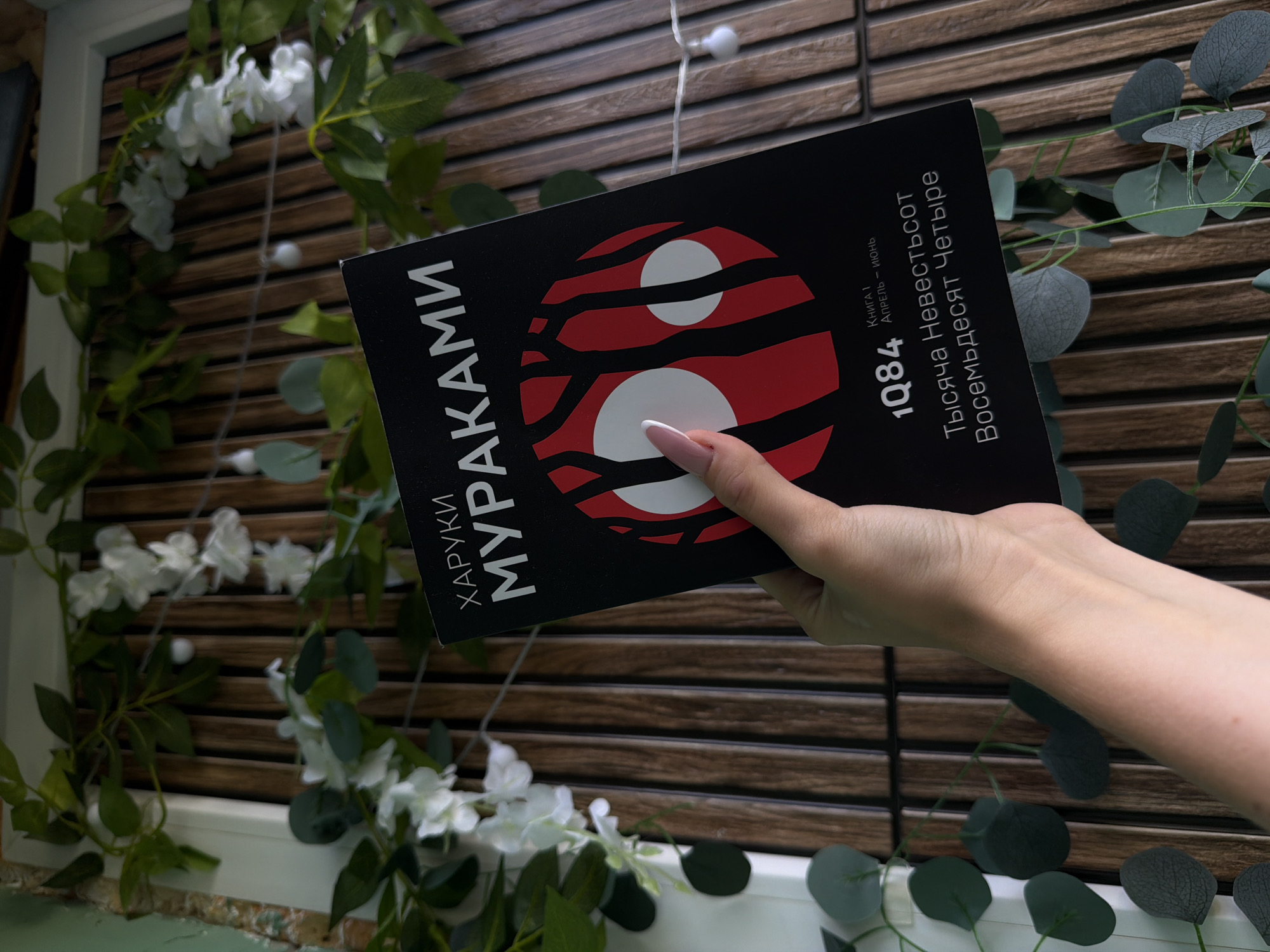 A hand holds a black book with white lettering. The book is titled "1Q84" and features a graphic design with red and white circles and black shapes resembling trees. The background consists of vertical wooden slats, adorned with artificial green foliage and small white flowers. Some fairy lights are visible strung across the background. The scene appears to be indoors or on a covered patio, with the wooden slats suggesting a decorative element in a home or a retail space. There is no discernible location in the city of Domodedovo, Russia, mentioned or suggested by the image.