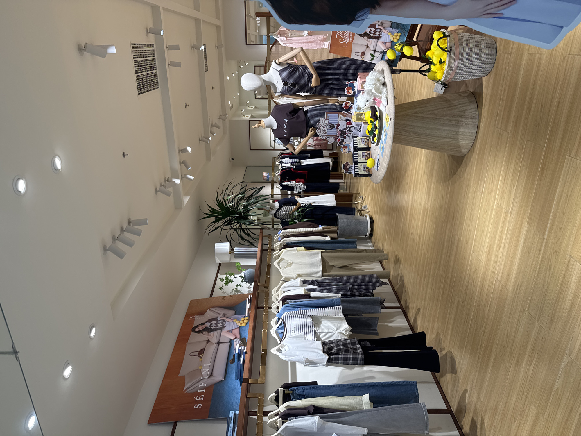 The image shows the interior of a well-lit clothing store, likely located in Linghe, China. The store features a wooden floor and white walls, with clothing racks displaying a variety of women's apparel. In the foreground, several mannequins are positioned around a circular display table adorned with decorative items and what appear to be lemons. To the left, a large banner with a fashion advertisement is mounted on the wall. The ceiling is equipped with multiple recessed and track lighting fixtures. The overall impression is of a modern retail environment.