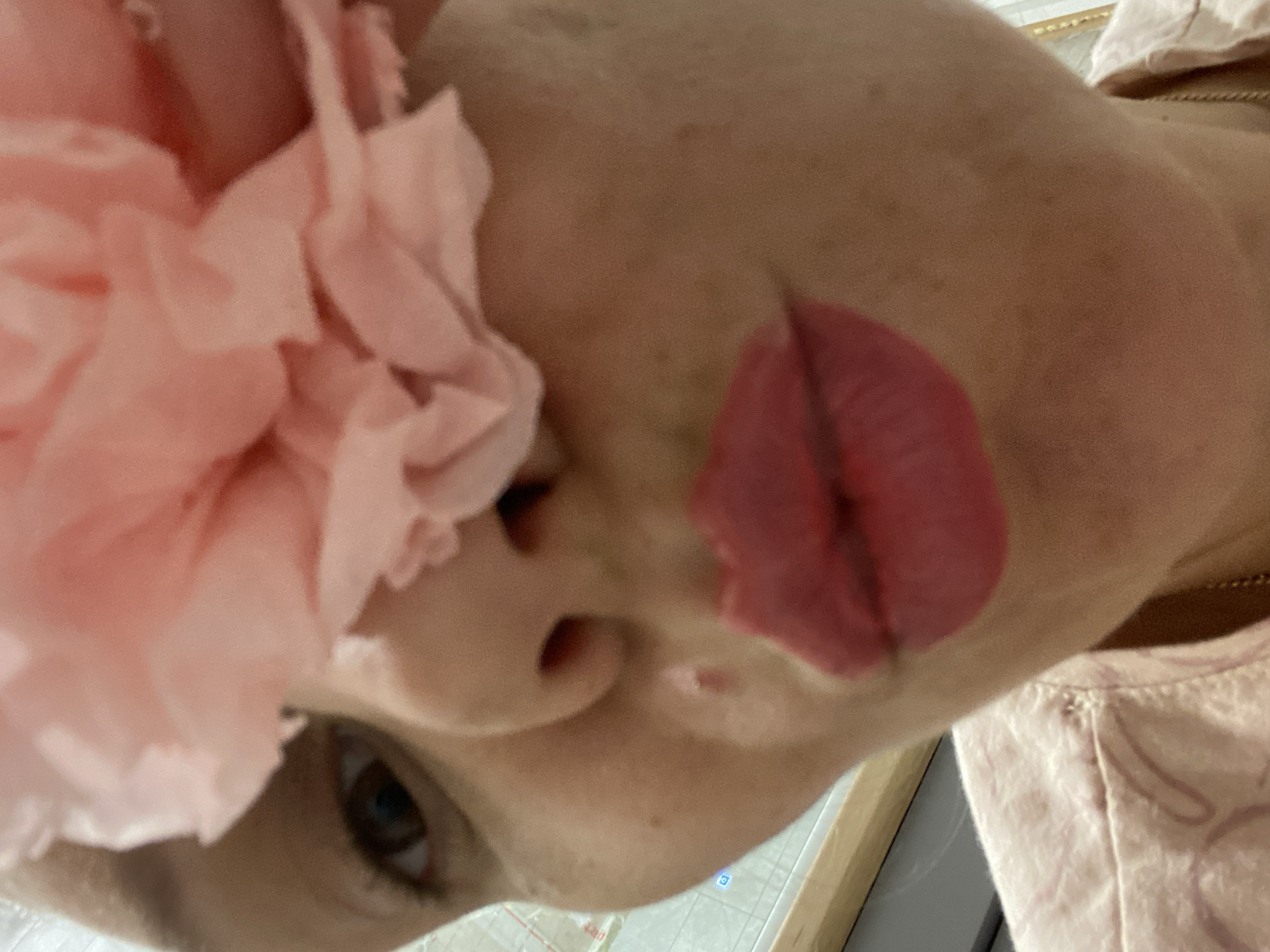 A close-up, tilted view of a person's face with a large pink paper flower obscuring the upper left area. The person has brown eyes, and fair skin. The lips are prominently visible and colored pinkish-red. A small portion of a light pink shirt is visible in the upper right and lower portion of the image. A wooden surface is also visible in the lower right. The location is not discernible from the image.