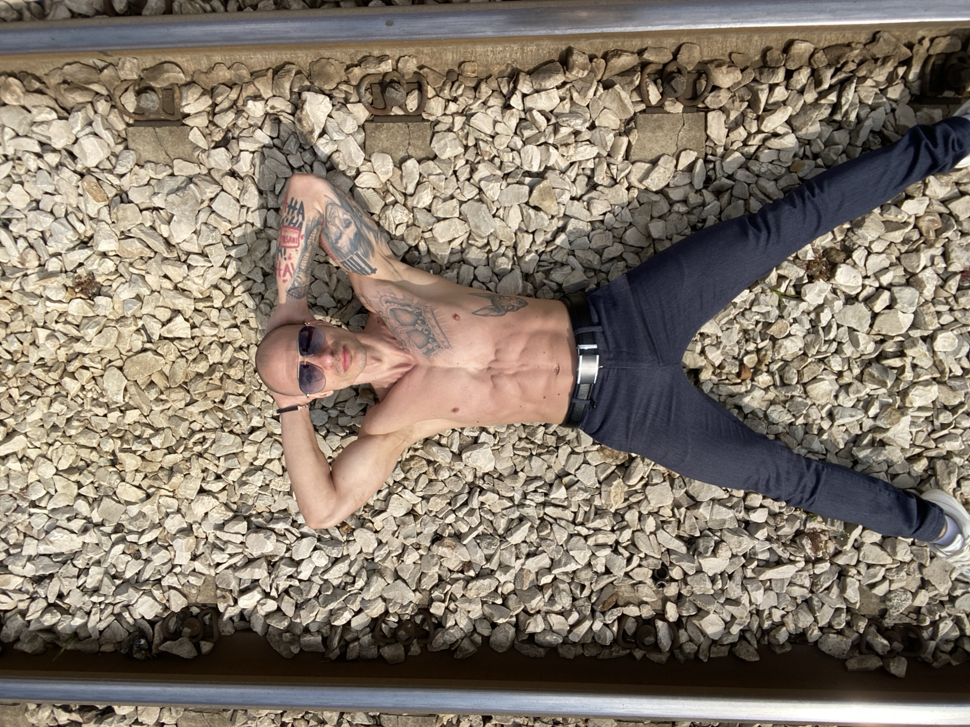 A shirtless man is lying on his back on a railroad track composed of gravel and metal rails. He is wearing jeans, a belt, sunglasses, and a bracelet on his left wrist. The man has a shaved head and numerous tattoos on his arms, chest, and abdomen. The location appears to be an outdoor railroad track. It is unknown if this is located in Pleven, Bulgaria.