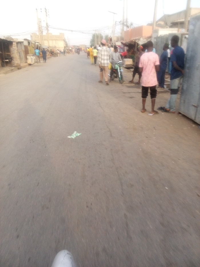 A straight asphalt road runs through a town. On both sides, various people are walking and standing. One man is on a small motorcycle. Some buildings and shops line the edges of the road, with utility poles visible in the distance. Litter lies on the road. The overall lighting suggests either early morning or late afternoon.
