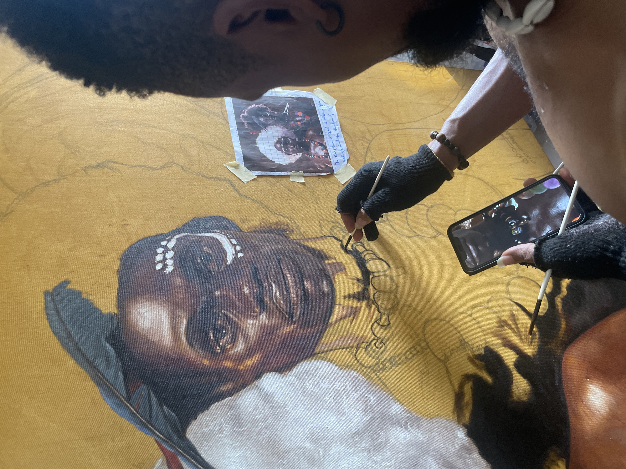 A close-up, high-angle indoor shot shows a person painting on a large canvas. The person is likely in Ado, Nigeria, but this is not evident from the photograph itself. The canvas is mustard yellow and features a partially completed portrait of a Black individual with distinct facial features, including a prominent nose, dark eyes, and full lips. The individual in the portrait wears white ornamentation on their head and a white garment. Feathers are visible as part of the portrait.
The artist is positioned above and to the side of the canvas. They have dark curly hair and are wearing an earring. They are using a thin paintbrush to add details to the painting, seemingly following a grid-marked photograph taped to the canvas above the portrait. A smartphone displaying an image resembling the painting's subject is held in the artist's other hand. The artist wears black fingerless gloves on both hands. A beaded bracelet adorns one wrist.
The painting is at an intermediate stage, with the face largely completed and outlining details sketched for the surrounding elements. The environment appears to be a studio or indoor workspace, with the canvas laid horizontally.