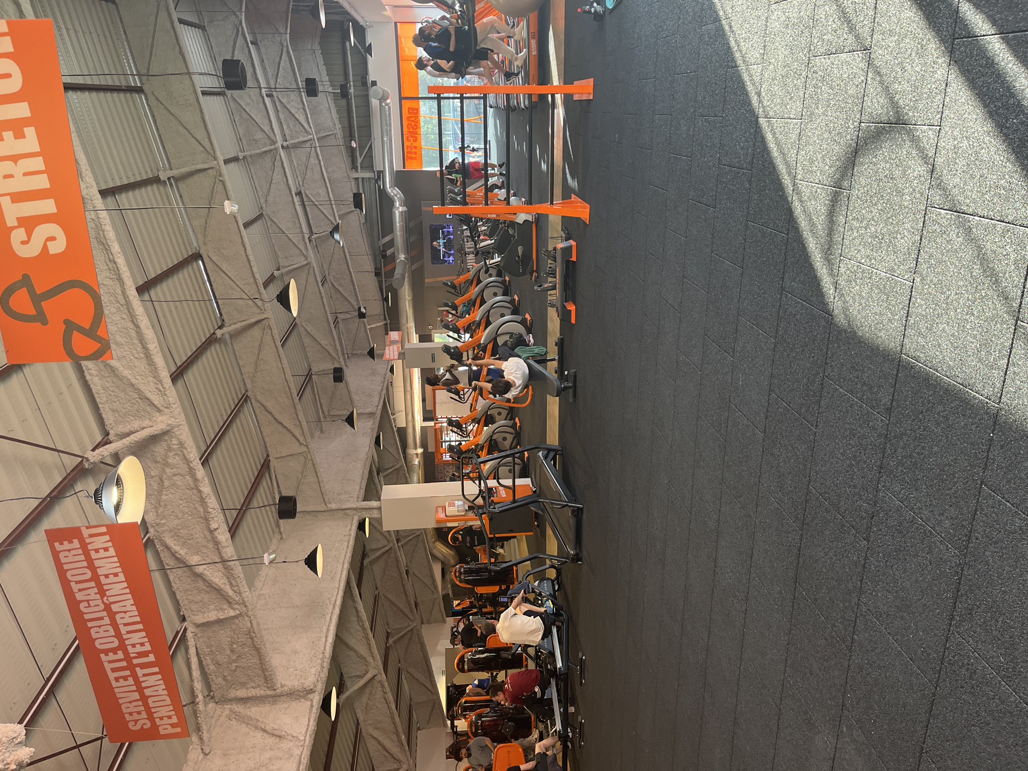 The image depicts the interior of a Basic-Fit gym in Illkirch-Graffenstaden, France. Rows of orange exercise bikes are occupied by individuals engaged in workouts. Overhead signage in French is visible, including one for "STRETCH" and another stating "Serviette Obligatoire Pendant L'entrainement" (Towel Required During Training). The gym features a high ceiling with visible support beams and ventilation. The floor is a dark, textured material. Sunlight streams in from windows at the far end of the gym.