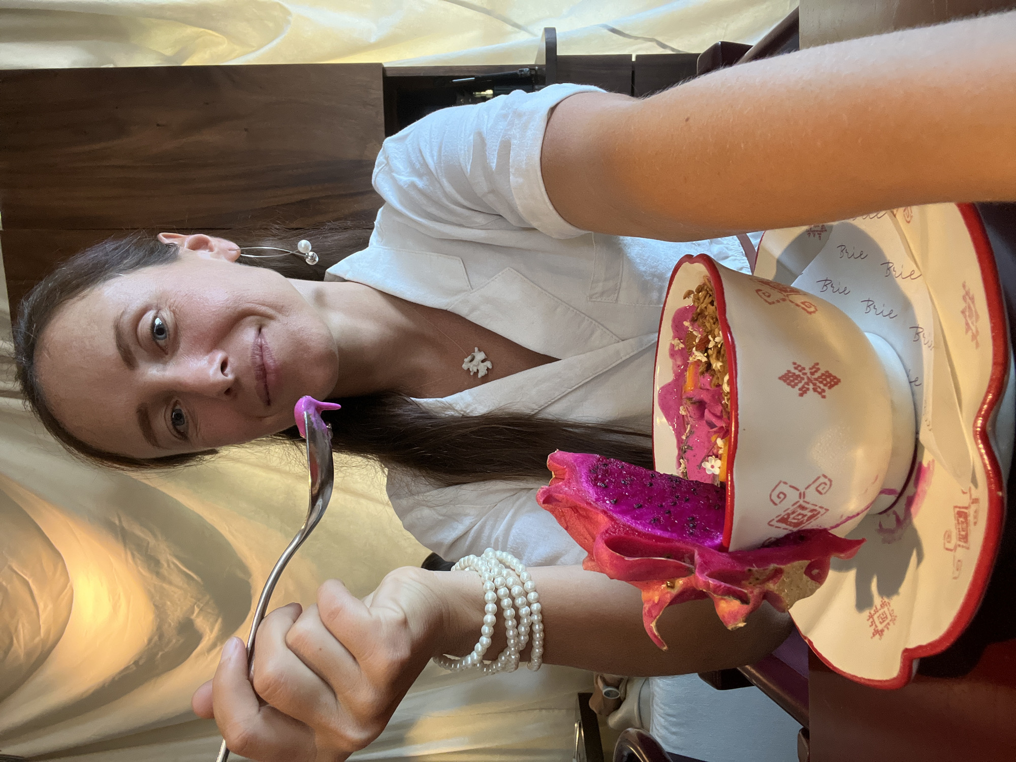 A woman in a white shirt is holding a spoon with pink dragon fruit yogurt and a piece of dragon fruit. The food is in a decorative bowl, which rests on a scalloped saucer. The woman is wearing pearl bracelets and a flower-shaped necklace. Her left earring consists of a silver hoop with two pearls. The scene appears to be indoors, possibly at a restaurant in Ubud, Indonesia, given the backdrop which includes draped fabric and dark wood paneling.