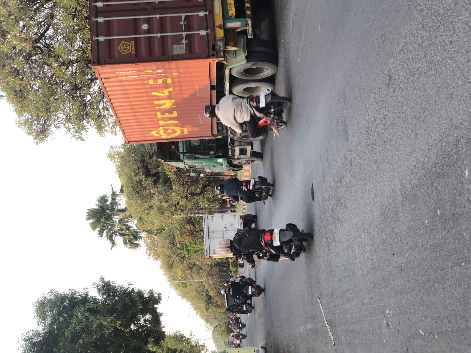 The image captures a street scene in Ngaban, Indonesia, featuring a busy road with several motorcycles and a large container truck. In the foreground, a person is riding a motorcycle, with a large backpack. Behind them, another motorcycle is ridden by a person wearing a helmet. Further down the road, more motorcycles are visible, indicating moderate traffic. The dominant object in the scene is a large, orange container truck with "TEMAS" written in yellow lettering. The truck is partially obscuring a white box truck. The environment appears to be a city street lined with trees, with a bright sky in the background. The lighting suggests it is either early morning or late afternoon.