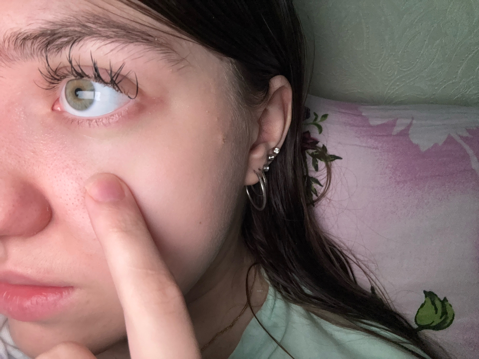 The image is a close-up, indoor portrait of a young Caucasian female, positioned in Zavolzhye, Russia. She is looking to her left, with her left eye and cheekbone prominently featured. Her skin appears to have visible pores on the cheek. Her left eye is light green with thick, dark eyelashes. She has dark brown hair that falls around her face and shoulders. Her left ear is visible, adorned with two small silver stud earrings and a larger silver hoop earring. A fingertip from her right hand is touching her left cheek near the nose. She is wearing a light green top. The background shows a pillow with a pink and white floral pattern, and a light green textured wall. The lighting is soft and appears to be natural or from an indoor source.