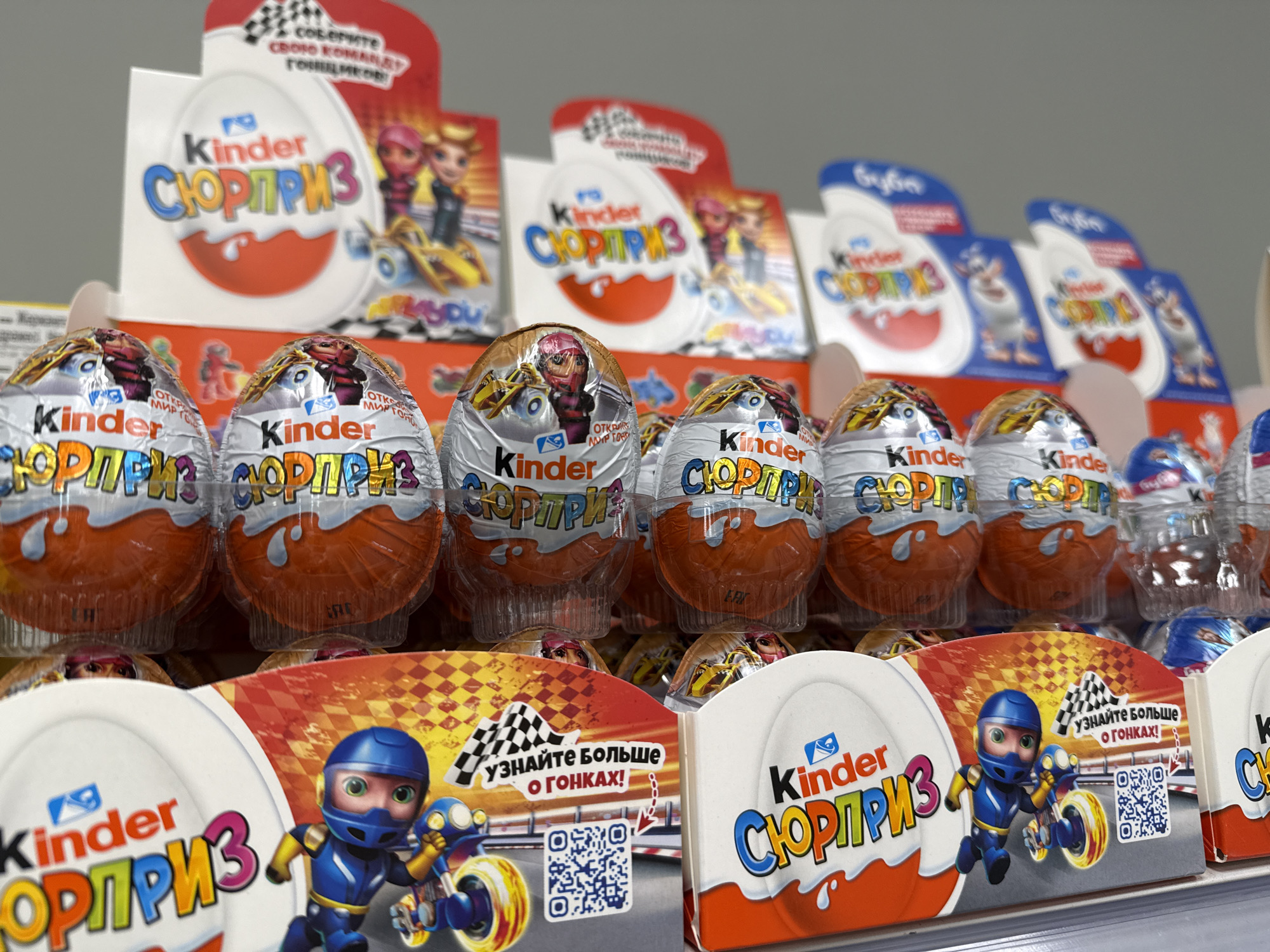 The image shows a shelf stocked with Kinder Surprise eggs, particularly the "СЮРПРИЗ" variety, indicating a Russian market presence. The eggs are prominently displayed, with special edition packaging featuring racing themes with characters and checkered flag motifs. Point of sale displays for the same product line, highlighting racing elements, are positioned in front of and behind the eggs. The overall scene appears to be a retail setting, likely a supermarket aisle or checkout area, in the city of Unknown, Unknown.