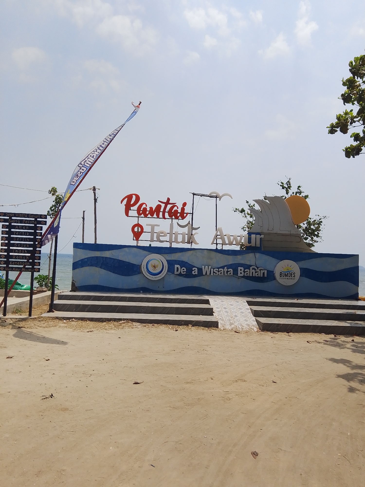 The image shows the entrance to Pantai Teluk Awur, a beach resort located in the city of Pejagan, Indonesia. The entrance features a large, colorful sign with the words "Pantai Teluk Awur" in red and white lettering. Behind the sign, there is a blue and white mural depicting waves and a sunset. To the left of the sign, a flag with the Indonesian national colors of red and white flies in the breeze. A sandy path leads up to the entrance, and trees and vegetation can be seen in the background. The sky is clear and blue, with a few white clouds. The overall scene conveys a sense of relaxation and vacation.