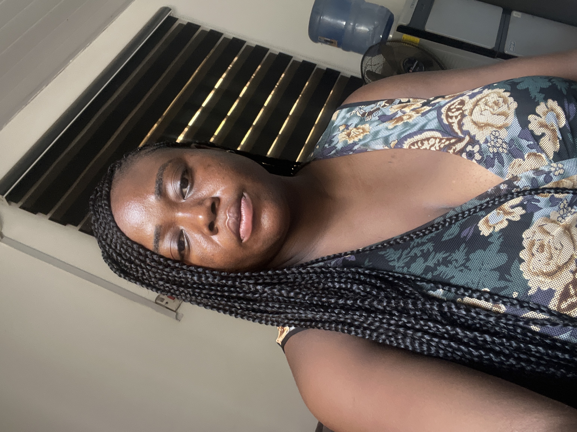 An indoor portrait captures a person, likely a woman, with dark skin and long black braided hair, looking directly towards the camera with a calm, soft expression. She is wearing a dark-colored top adorned with a floral pattern featuring motifs in blue, green, and beige/gold.
The setting appears to be a room, possibly a home or office. In the background, a window with horizontal blinds is visible, allowing streaks of sunlight to penetrate, indicating it is daytime. To the person's right, a large blue water dispenser bottle and a black electric fan are discernable, alongside other indistinct objects on a surface. The wall in the background is a light, neutral color.
The overall impression is a casual, close-up shot. The presence of a fan and water dispenser aligns with typical household items in warmer climates, consistent with the provided location context of Abuja, Nigeria. No visible text is present in the image.
