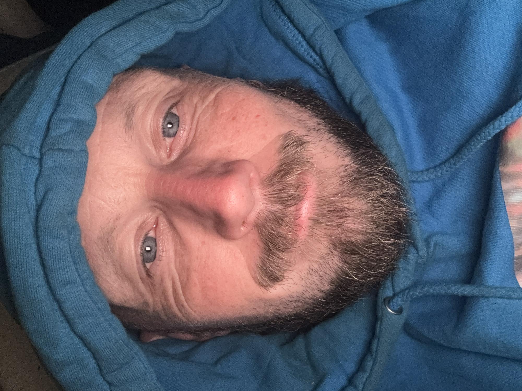 The image is a close-up, selfie-style shot of a man's face. He is wearing a blue hooded sweatshirt, with the hood pulled up around his head, framing his face. The man has blue eyes, fair skin with some redness on his cheeks and nose, and a goatee and mustache. He appears to be looking directly at the camera with a neutral expression. The background is dark and nondescript. There are no identifiable landmarks or environmental clues within the image that would allow for pinpointing a specific location within Cheltenham, United Kingdom. The focus is entirely on the man's face and the blue hoodie.