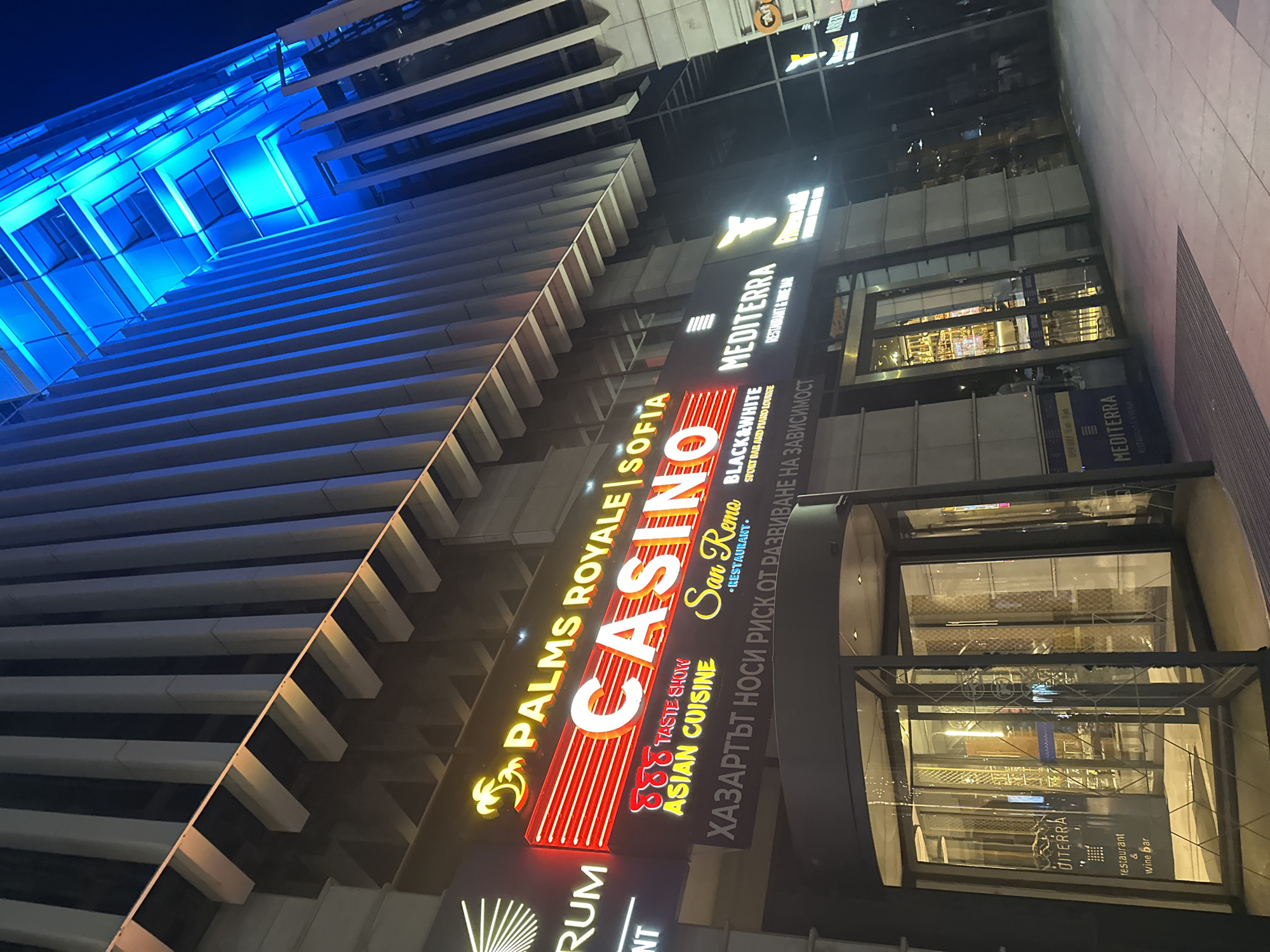 The image shows the exterior of a modern building at night, illuminated by various lights. The building's facade is characterized by vertical fins and is lit with bright blue lights at the top. A prominent neon sign advertises "PALMS ROYALE SOFIA CASINO", with smaller signs for "TASTE SHOW ASIAN CUISINE", "San Remo RESTAURANT", and "BLACK & WHITE SPORT BAR AND PIANO LOUNGE". Below these, in Cyrillic script, is a warning: "ХАЗАРТЪТ НОСИ РИСК ОТ РАЗВИВАНЕ НА ЗАВИСИМОСТ" (Gambling carries the risk of developing addiction). To the left of the casino sign, there's a partially visible sign for "RUM". To the right, signs for "MEDITERRA RESTAURANT & WINE BAR" and "Princess Bar" are visible. The ground level features a revolving glass door entrance to a establishment labeled "MEDITERRA restaurant & wine bar". The pavement in front of the building is visible, along with street lighting and shadows cast by the building. The scene appears to be a commercial district in Sofia, Bulgaria, given the signage.