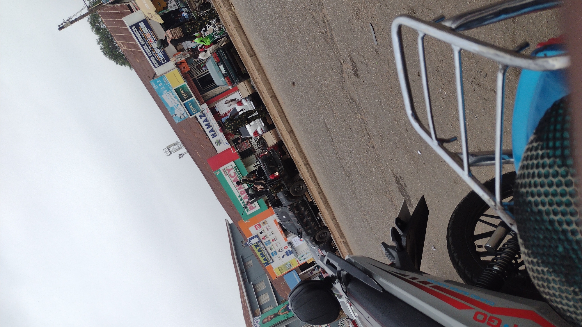 This image captures a bustling street scene in an urban environment, likely in Jos, Nigeria, on an overcast day. The photograph is taken from the perspective of someone on a motorcycle, with parts of the vehicle, including a metallic cargo rack and a wheel, visible in the foreground on the right. The motorcycle itself has "GO" emblazoned on its side.
The left side of the frame features a row of commercial buildings with corrugated iron roofs, housing various businesses. Their facades are adorned with numerous colorful signs and advertisements. The street is paved, and several vehicles are present, including cars, a dark-colored utility vehicle or truck, and other motorcycles parked or moving along the road.
People are visible throughout the scene, engaged in daily activities. Some individuals are seen wearing camouflage uniforms, suggesting the presence of security personnel, while others are civilians walking or standing near the shops.
Notable text on the signs includes:
* "OLUWA GLOBAL RESOURCES (TONERO COMMUNICATIONS)" with a phone number (e.g., "00033842000").
* "HAMAZ COMMUNICATIONS" appearing on multiple storefronts.
* "NASARA PHARMACY".
* Logos for "DStv" and "Gotv", indicating satellite television services.
* Other smaller, less legible text also appears on various advertisements.
The overall scene depicts a vibrant commercial area characterized by local businesses and everyday urban life.