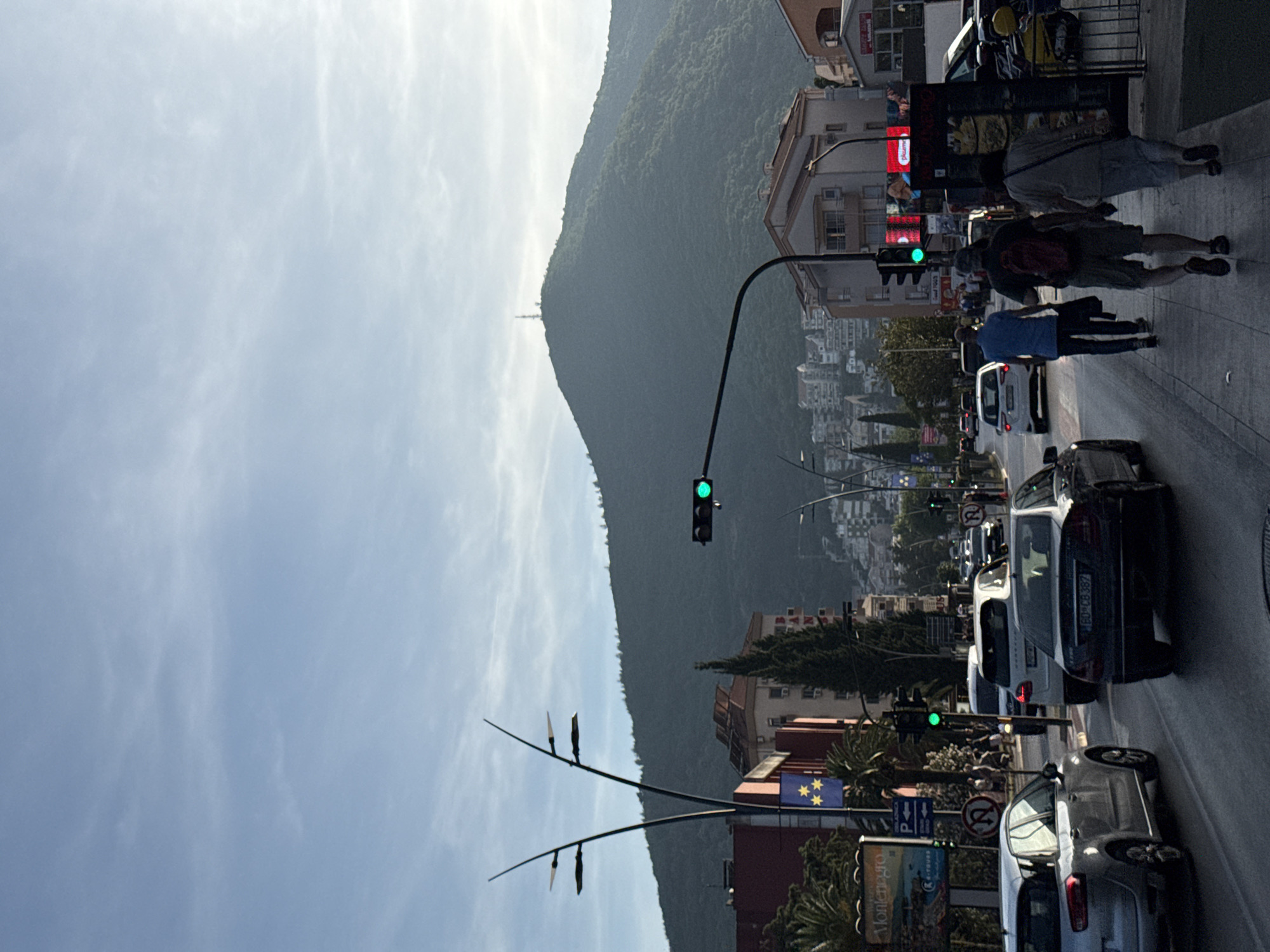 This image presents an active street scene in a coastal town, likely Budva, Montenegro, as suggested by visible cues and context. A prominent, heavily forested mountain dominates the background, rising steeply under a bright, slightly cloudy sky. A small tower or structure is visible near the mountain's peak.
The foreground features a multi-lane road with several cars in traffic, indicating a busy period. One car's license plate, "BD 08-387," explicitly points to Budva (BD is the vehicle code for Budva). Multiple traffic lights, showing green, regulate the flow of vehicles. On the right side, a sidewalk is populated with pedestrians; two individuals in the immediate foreground are walking away from the viewer, one with a red backpack and another in a blue shirt. More people can be seen further along the sidewalk.
Buildings of various sizes, housing businesses and residences, line the street. Street lamps with multiple light fixtures are spaced along the road. A flag, yellow with blue stars, is mounted on a pole on the left side, possibly representing a local municipality or region. Below it, a large sign or billboard clearly displays the word "Montenegro" alongside a scenic photograph. Other road signs, including a "no U-turn" symbol, are also visible.
The scene appears to be set during daytime, possibly late afternoon, with fair weather and good visibility, capturing the everyday hustle and bustle of an urban area nestled against a dramatic natural backdrop.