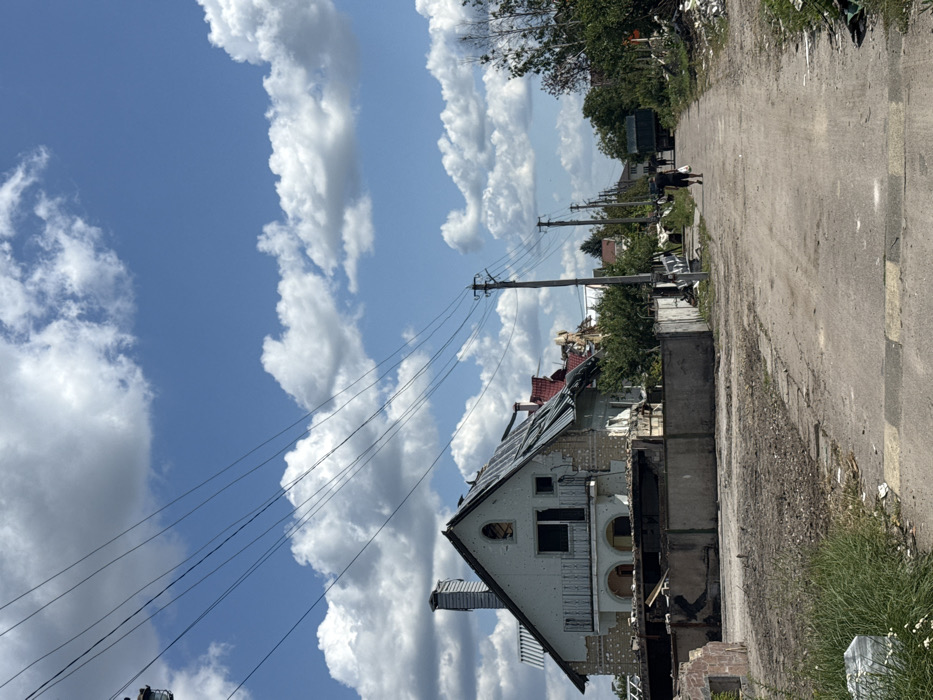 The image captures a residential street in Markhalivka, Ukraine, under a clear blue sky dotted with scattered white cumulus clouds. The scene exhibits significant structural damage to buildings. On the left side, a two-story house with a light-colored facade and a grey, pitched roof is prominent. Its roof shows extensive damage with large sections of material torn and missing, exposing underlying structures. The house's facade is perforated with numerous small circular impacts and larger breaches, particularly near the ground level, and its balcony railing is partially dislodged. A damaged concrete fence lies in front of this property, with debris scattered on the ground. Further down the street on the left, another building with a red roof appears heavily damaged or partially collapsed.
A unpaved, dirt and gravel road, with some segmented paved sections, extends into the distance, leading towards a visible large, dark-colored vehicle, possibly a truck. Utility poles with overhead power lines run along both sides of the road, receding into the background. The right side of the road is lined with dense green vegetation, including trees and bushes, intermingled with debris and rubble. Two individuals are walking away from the camera down the road; the closer figure, dressed in dark clothing (shorts and a t-shirt), carries a white bag in their right hand. The immediate foreground shows a rough, unpaved surface with patches of grass and scattered discarded items, including a white object in the lower left.