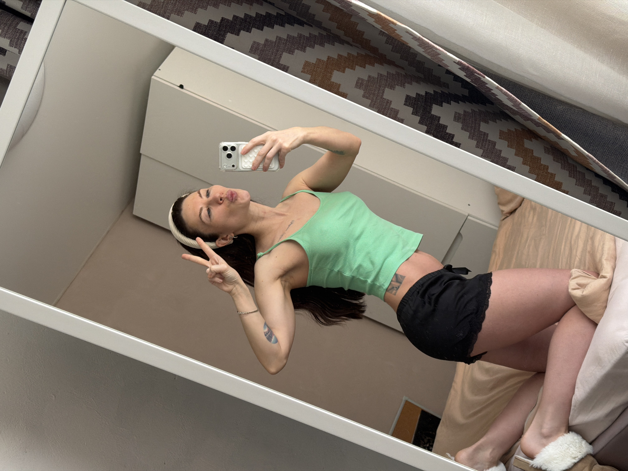 A woman is posing for a selfie in a mirror. She is wearing a green tank top and black shorts. Her hair is dark. She is holding a smartphone in her right hand and making a peace sign with her left hand. The reflection shows a bedroom with a white dresser and a bed with patterned curtains. The location is most likely not Perm, Russia, as there is no indication in the image.
