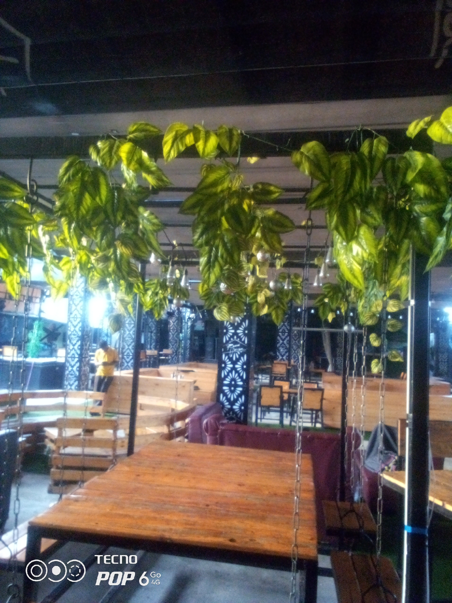 This is a medium shot, indoor photograph taken at a restaurant in Howayi, Nigeria. The scene is decorated with hanging plants and chain swings, giving it a rustic and natural ambiance. In the background, a person wearing a yellow shirt is standing near a staircase, possibly an employee or patron. Several wooden tables and chairs are arranged throughout the space, suggesting it's a dining area. The lighting appears to be artificial, with some hanging lamps visible, contributing to the establishment's atmosphere. The image's lower-left corner displays a "TECNO POP 6 4G" watermark, indicating the device used for capture.