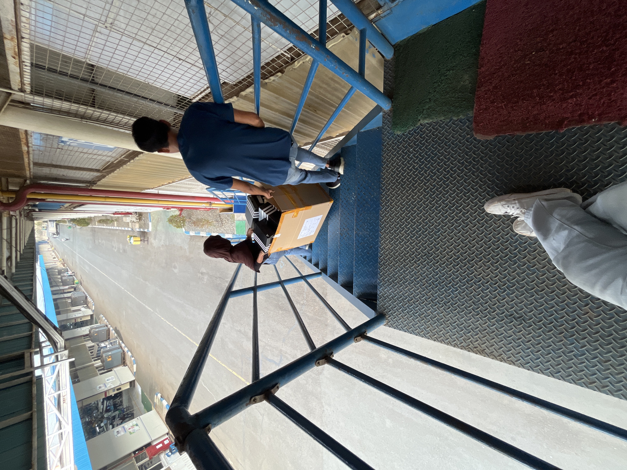 The image depicts an outdoor industrial setting, possibly a factory or warehouse complex, with elements of infrastructure visible, including a long building with blue accents and what appears to be a road or walkway. Two individuals are ascending a metal staircase with blue railings. The person in front, wearing a dark hooded garment and jeans, is carrying a cardboard box filled with black shoe boxes. Behind them, a person in a blue t-shirt and jeans is also on the stairs. The staircase is constructed from diamond plate metal on the treads and has a non-slip green mat at the bottom. The perspective is from a higher vantage point, looking down the stairs and across the industrial area. The specific location in Pelang, Indonesia, cannot be determined from the visual information alone.