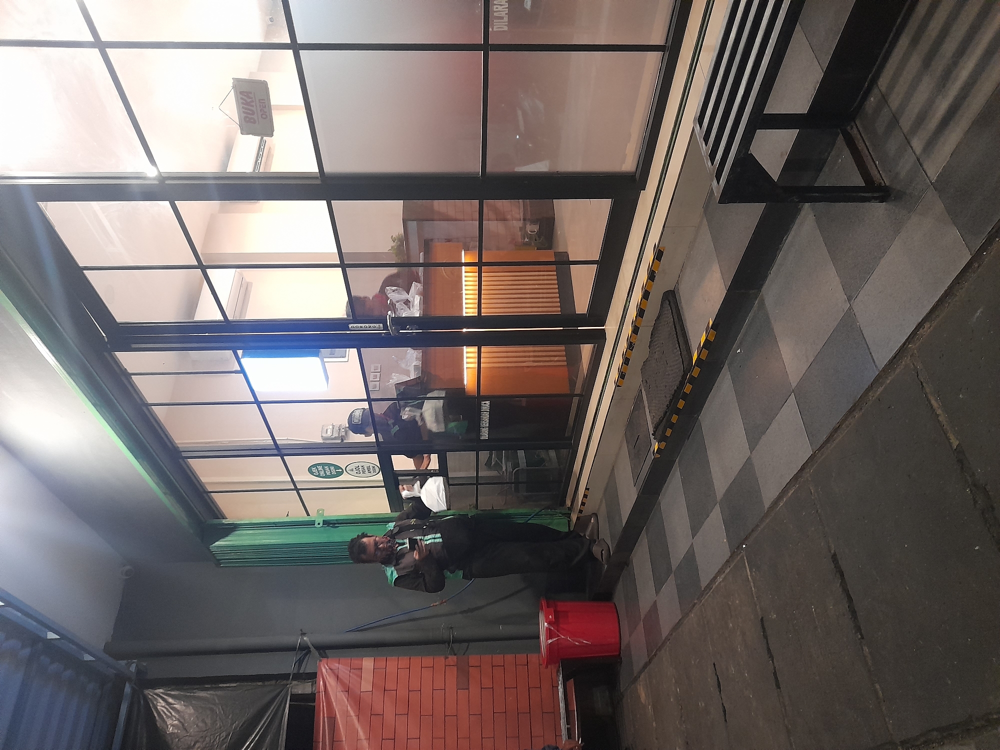 A man wearing a dark jacket with a teal accent and dark pants is standing outside a building with large glass windows. He is looking down at his phone and holding a white plastic bag in his left hand. The building's interior is visible through the windows, showing a counter with several people behind it and some white bags on the counter. A small sign hanging in the window reads "BUKA OPEN". The exterior area features a red trash can, a tiled floor with a checkered pattern, and a dark gray paved area. A black metal bench is partially visible to the right. The overall scene suggests a commercial establishment, possibly a food vendor or restaurant, in North Jakarta, Indonesia, with the man appearing to be a delivery person.