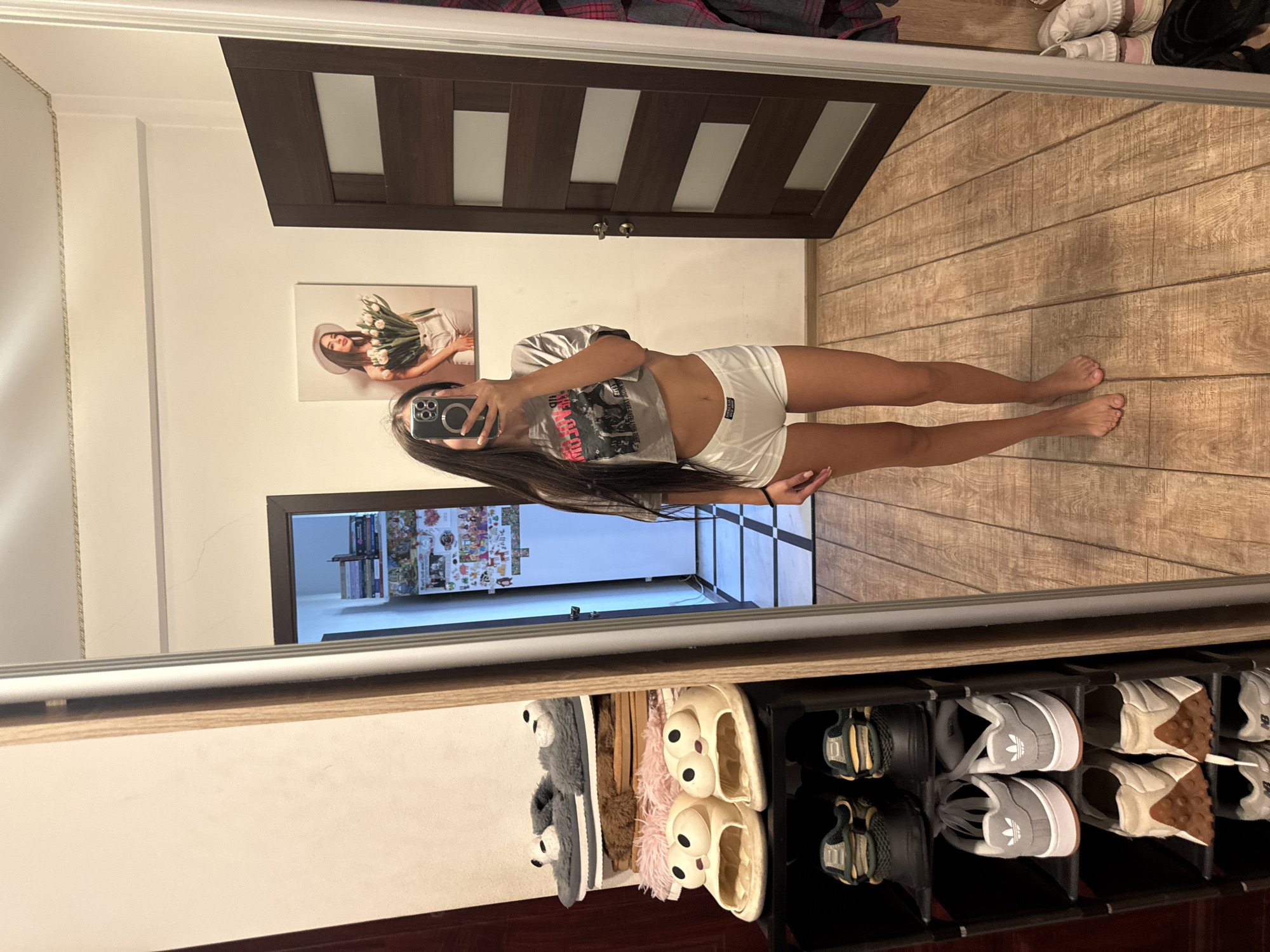 A full-body, vertical, mirror selfie captures a young woman standing in a room. She is wearing a gray t-shirt with a graphic print and white shorts. Her long, dark hair falls down her back. In the background, a large wooden door with vertical panels is visible. To the left of the door, a framed picture hangs on the wall, depicting a woman wearing a hat and holding a bouquet of white tulips. The floor is made of wooden planks. In the lower portion of the image, a shoe rack is visible, filled with various pairs of footwear, including slippers with large googly eyes and sneakers. The lighting suggests it is daytime. The scene appears to be an indoor residential setting, likely a bedroom or dressing area. There is no visible text.