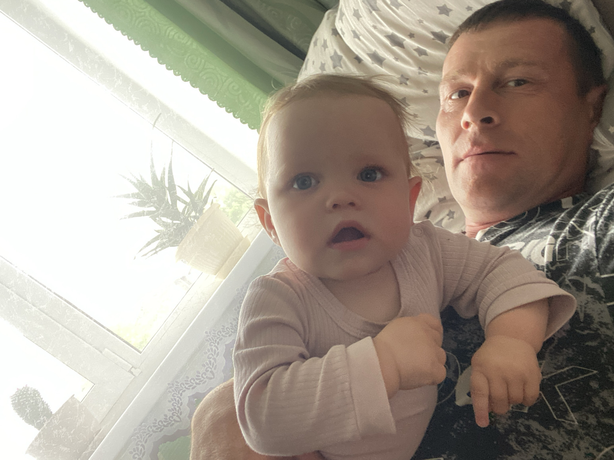 This close-up indoor image features a man and a baby, likely taken as a selfie. The man, with short dark hair and a neutral expression, is lying down, possibly on a bed covered with white sheets patterned with grey stars. He is wearing a dark t-shirt with a subtle pattern. The baby, with light hair and blue eyes, is positioned on or next to the man, looking directly at the camera with a wide-eyed, slightly surprised expression and an open mouth. The baby is dressed in a light pink, ribbed long-sleeved onesie.
The bright illumination from a window on the left side of the frame indicates it is daytime. Through the window, green foliage and potted plants are visible; one appears to be an aloe vera plant, and another plant resembling a cactus is partially seen. Green curtains with a scalloped, decorative pattern are visible at the top left of the window, while a white curtain with a lace-like pattern is on the right. The scene conveys a moment of domestic closeness, set in what appears to be a bedroom in Фоки, Russia. No text is visible in the image.