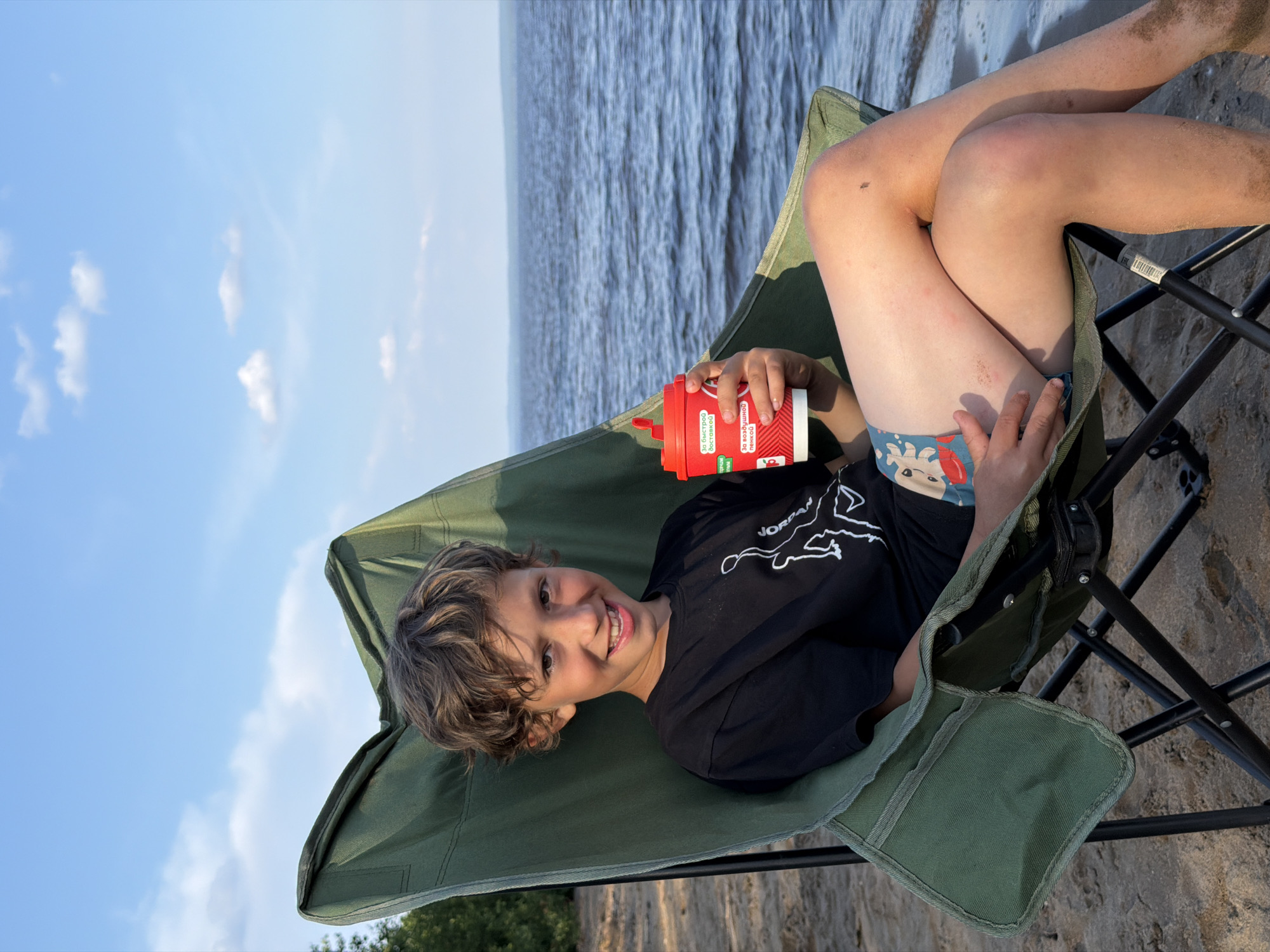 The media file shows a young boy sitting in a green folding chair on a sandy beach. He is looking towards the camera with a smile, his head tilted slightly. The boy has light brown, wavy hair and is wearing a black t-shirt with a white outline graphic of a figure. His legs are bent, and he is holding a red plastic cup with a white lid and straw in his right hand. The cup has some red and white text on it, including what appears to be Russian writing. He is also wearing patterned shorts with images of what look like cartoon characters. The background consists of a large body of water, likely a river or lake, with ripples on the surface, and a blue sky with scattered white clouds. The lighting suggests it is daytime, possibly late afternoon, given the angle of the sun casting shadows. There are small insects visible on the boy's legs and the sand. The setting appears to be a natural environment, likely a recreational area near water.