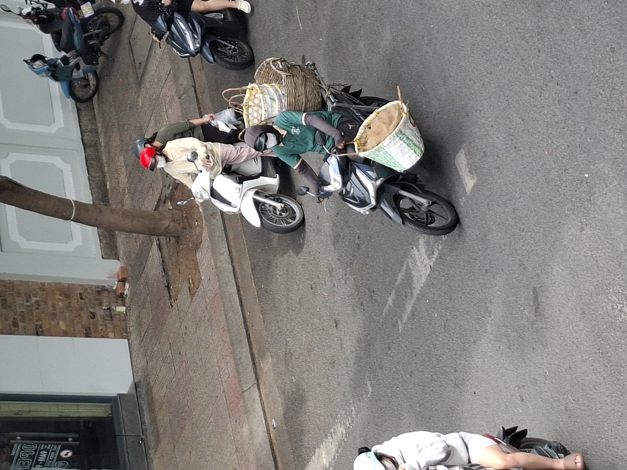 In Nha Trang, Vietnam, on a paved city street, a scene unfolds with several people on motorbikes. In the foreground, a person wearing a green shirt, grey arm sleeves, a hood, and a white face mask rides a dark-colored motorbike. They are carrying two large woven baskets, one on the back of the motorbike filled with eggs, and another smaller woven basket hanging from the front, containing a tan material. To the left, another motorbike carries two individuals, one in a light-colored jacket and pink pants, wearing a red helmet and a white face mask, and the other in a green shirt, carrying a plastic bag. Further back, a blue motorbike is parked. The environment includes a sidewalk with brick paving and a building with a white facade. The lighting suggests it is daytime.