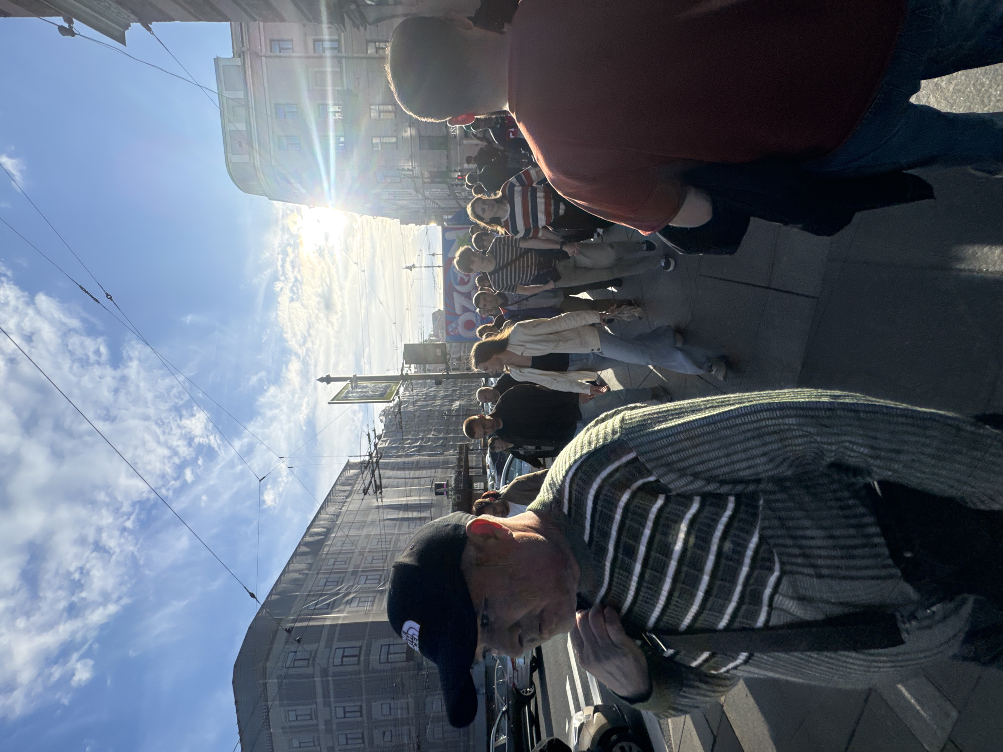 The image shows a group of people walking down a street in Saint Petersburg, Russia. The sun is shining brightly, and there are a few clouds in the sky, suggesting it is daytime. The people are dressed in casual clothing, and some appear to be looking at their phones. A building is visible in the background, and there are some wires overhead, possibly from a tram or trolleybus system, which are common in Saint Petersburg. The overall atmosphere seems relaxed, with no indication of any particular event. The image is oriented vertically, with the people walking from the top of the frame towards the bottom. The people in the foreground are out of focus, while the people further back are more in focus. There is no visible text in the image.