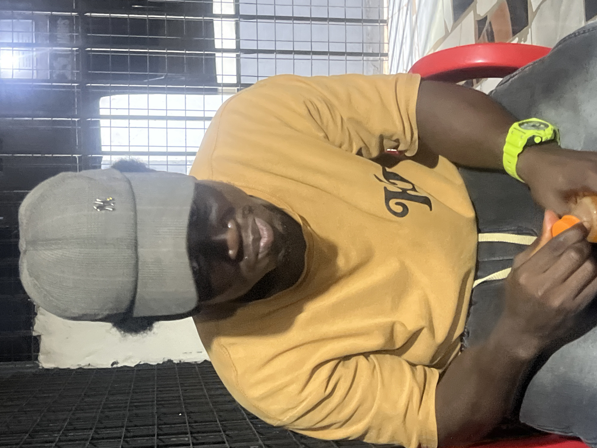 This media file shows a person in Nkpor, Nigeria. The subject is wearing a mustard yellow t-shirt and a gray baseball cap. They are also wearing a bright green digital watch on their left wrist. The person appears to be sitting and leaning back, with their head tilted upwards. Their mouth is slightly open, and they seem to be looking at something out of frame. A piece of an orange object, possibly a fruit, is visible in their right hand, suggesting they might be eating or holding it.
The background is somewhat blurred but appears to be an indoor or sheltered area with a metal grate or mesh structure in the foreground and possibly a tiled wall in the background. The lighting suggests it might be daytime, though the specific time of day is not clearly indicated. The overall mood is casual and relaxed. There is no visible text in the image.