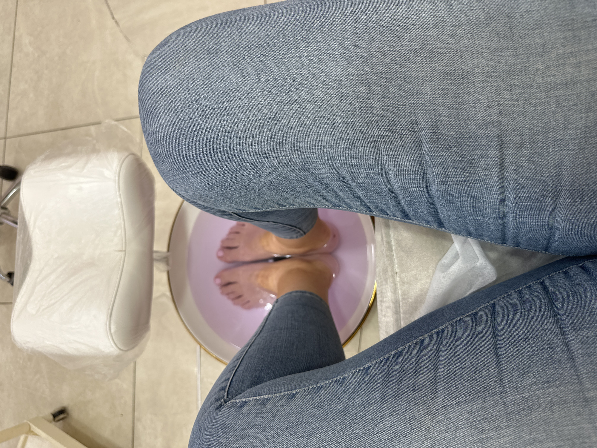 A top-down shot shows a person wearing light blue jeans sitting with their feet soaking in a round, shallow basin of light purple water. The basin has a gold rim. To the left of the basin is a white padded leg rest, partially covered in clear plastic. The floor beneath is beige tile. The location is likely a nail salon but the city is indeterminate.