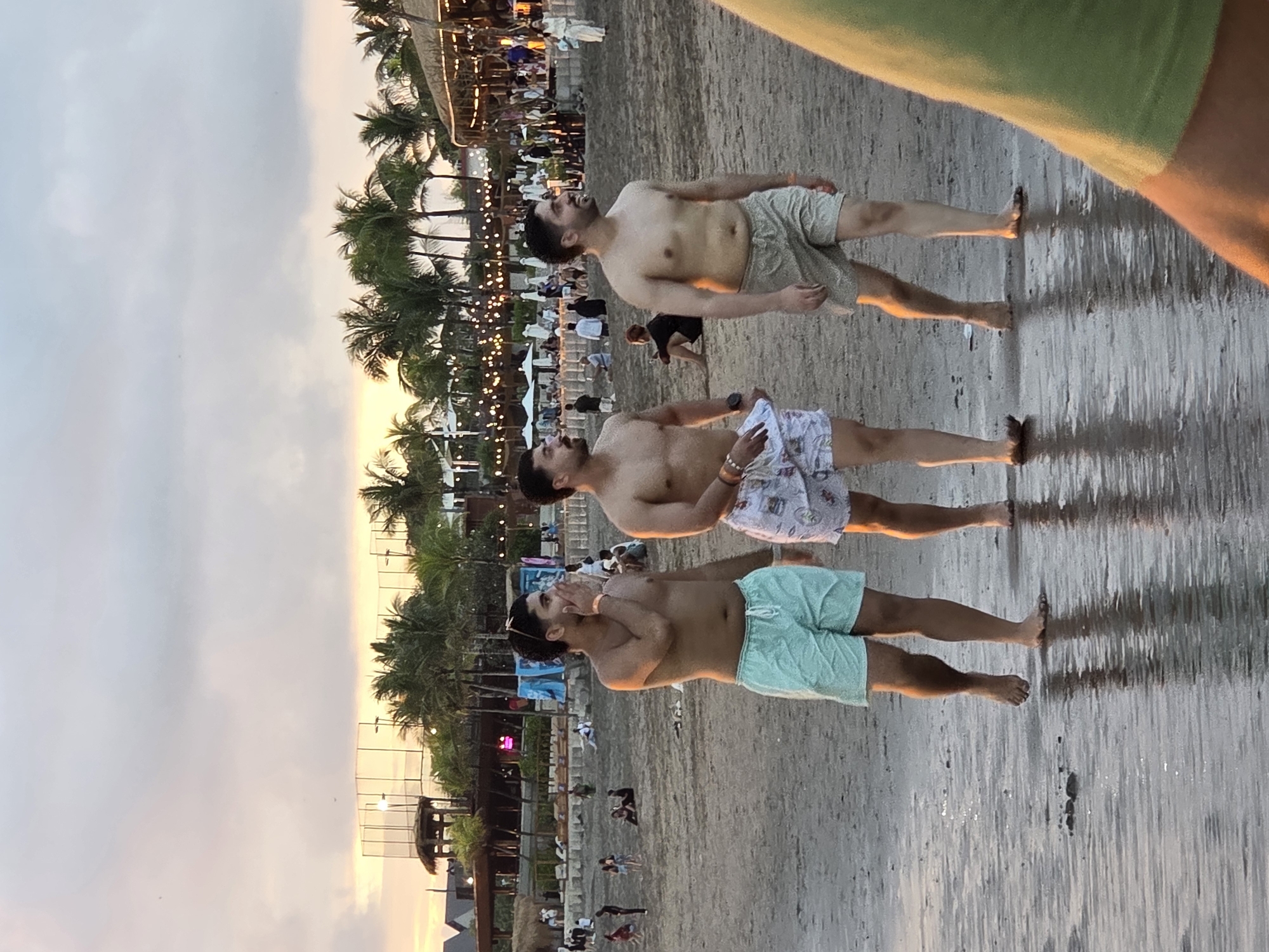 Three shirtless men in swim trunks walk along the wet sand of a beach at dusk. The man on the left wears light teal swim trunks, the middle man wears patterned swim trunks, and the man on the right wears light beige swim trunks. They are walking towards the right side of the frame. In the background, palm trees line the shore, with a building structure that appears to be a restaurant or bar with string lights visible. Other people are present on the beach in the distance, some sitting and others walking. The sky is overcast with soft hues of orange and yellow, indicating sunset. The location is identified as North Kuta, Indonesia.