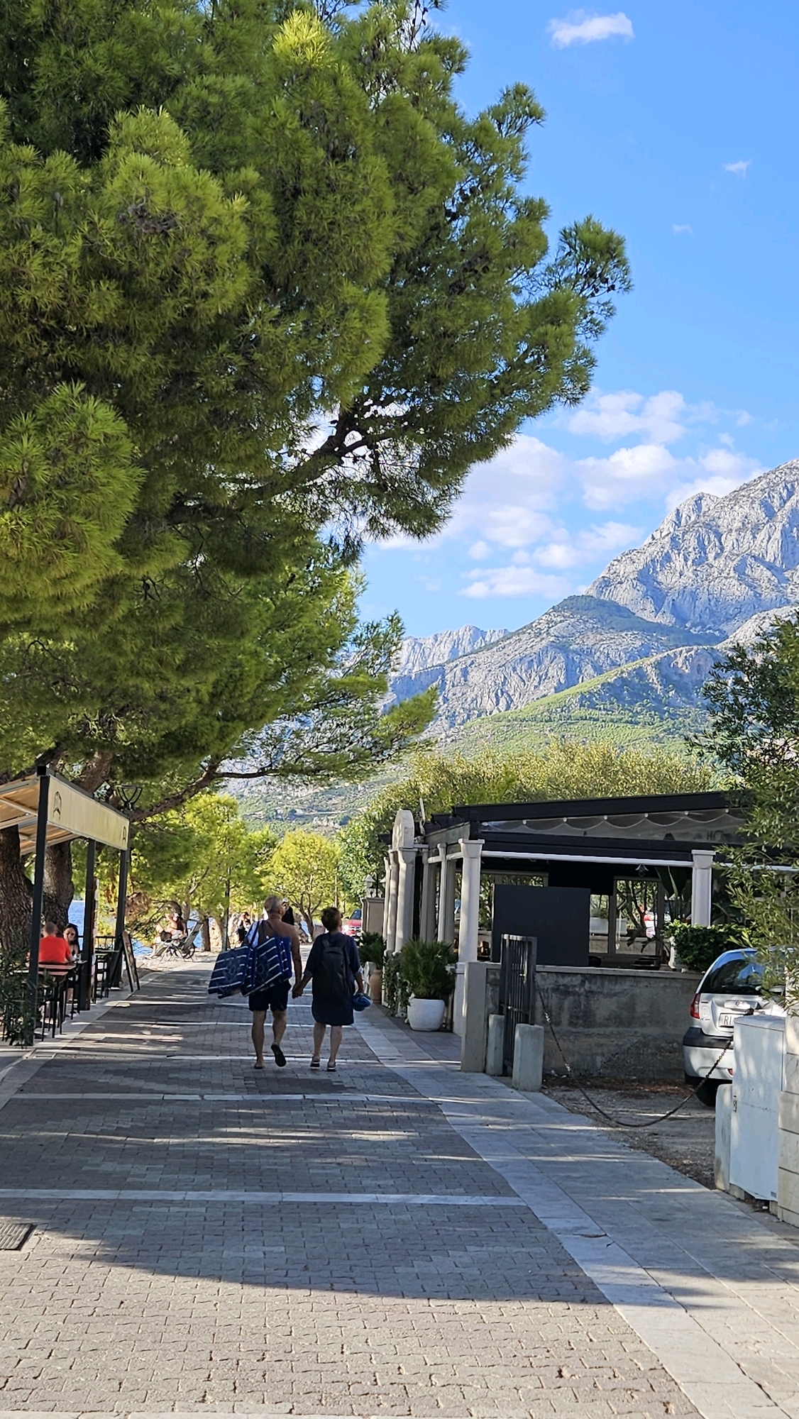 The image depicts a seaside promenade in Tučepi, Općina Tučepi, Croatia, on a sunny day. In the foreground, a paved walkway runs parallel to the sea, lined with trees on the left and a series of open-air establishments with white pillars on the right. A couple, a man and a woman, are walking away from the camera down the walkway. The man is carrying a striped beach towel over his shoulder. In the background, a mountain range with sparse vegetation dominates the horizon under a clear blue sky dotted with white clouds. The overall scene suggests a relaxed vacation atmosphere.