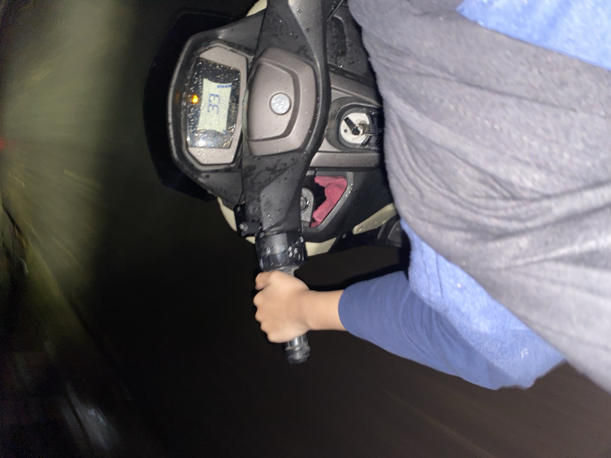 A person's hand, wearing a blue long-sleeved shirt, grips the handlebars of a scooter at night. The scooter's digital dashboard displays the number "33," suggesting a speed of 33 kilometers per hour or miles per hour. The environment is dark, with streaks of light indicating motion. Water droplets are visible on the dashboard, implying rain or high humidity. The background is blurred, obscuring any specific details of the location, but the presence of the road and surrounding darkness suggests an outdoor urban or suburban setting.