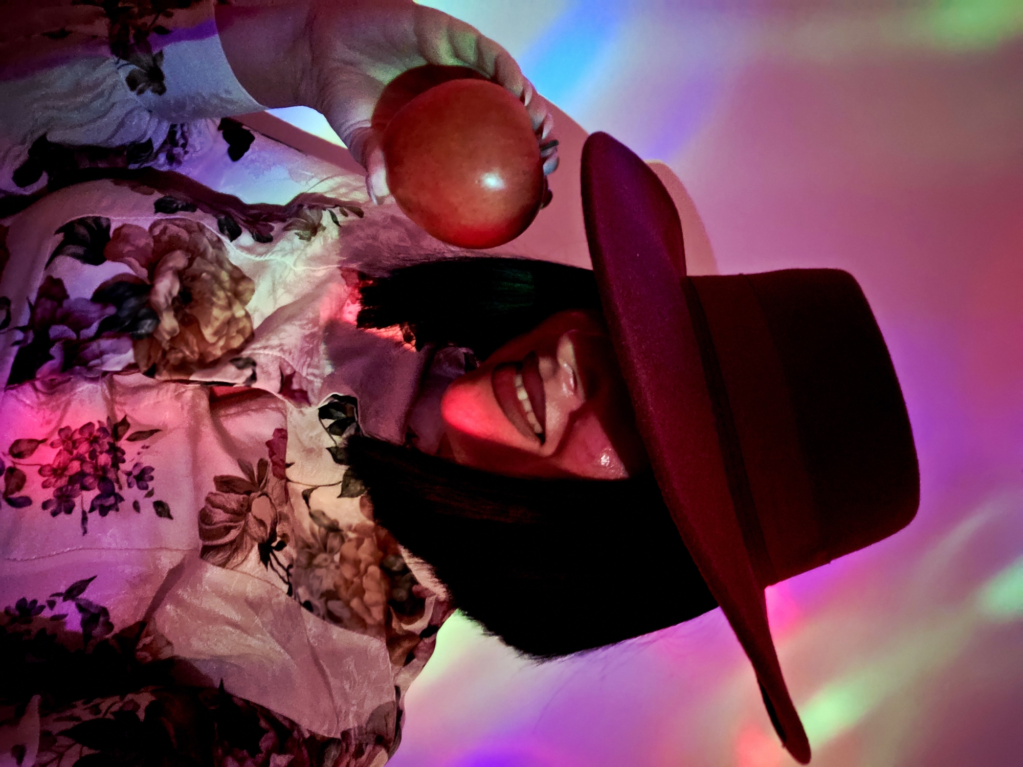 The image captures a highly stylized and vibrant portrait of a person, likely female, presented from an unconventional, tilted perspective. The central figure is shown holding a smooth, red apple in their left hand, which is adorned with light nail polish. Their face is partially obscured by a dark, broad-brimmed fedora-style hat.
A notable detail is the person's mouth, wide open to reveal white teeth and a pink tongue, conveying a sense of laughter, a playful shout, or an exuberant expression. Dark hair frames the face, adding contrast to the bright mouth. The person is wearing a garment with a prominent floral pattern, featuring large blossoms in shades of pink, maroon, and brown against a lighter fabric, visible on the left side of the frame.
The background is a dynamic wash of diffused, multicolored light, creating an atmospheric and surreal setting. Streaks and pools of fuchsia, purple, bright blue, red, and subtle green light illuminate the scene, suggesting artificial lighting such as projectors, stage lights, or perhaps a disco ball. There are no explicit location cues, but the lighting implies an indoor environment or a night-time creative shoot. The overall mood is energetic, playful, and artistic. No visible text is present in the image.