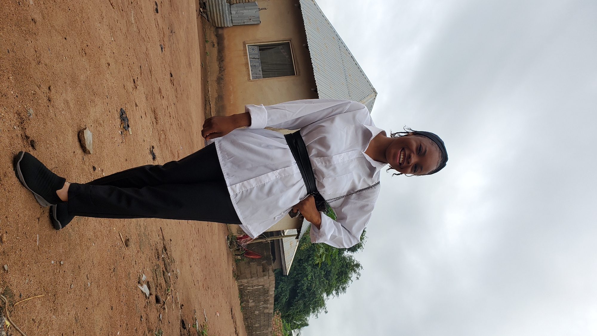 A young Black woman stands in front of a simple, light-colored building with a corrugated metal roof. She is wearing a white button-down shirt, cinched at the waist with a wide black belt, and black pants. Her hair is styled in braids and covered by a black headwrap. She is smiling broadly and looking at the camera, with her right hand on her hip and her left hand holding a black purse with a silver chain strap. The ground around her is sandy and appears to be a dirt road with some debris. In the background, there are more buildings and trees under a cloudy sky. The scene suggests a rural or semi-urban setting, consistent with areas in Jalingo, Nigeria.