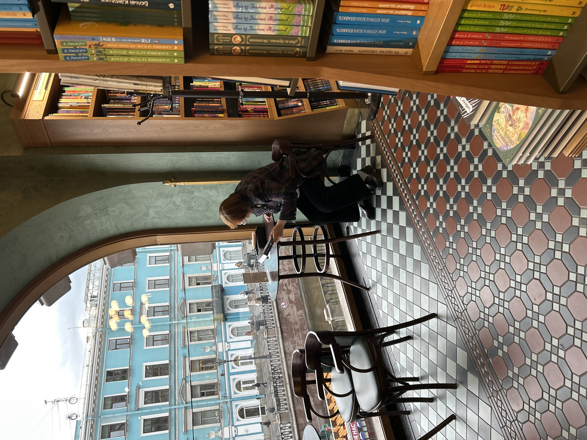 A photograph taken from an elevated perspective looking down, depicts the interior of a bookstore or café in Saint Petersburg, Russia, featuring a person seated at a table by a window. The individual, wearing a patterned shirt and dark pants, appears to be looking at something on the table. The window provides a view of the city outside, revealing a canal and a blue building with a white facade.
The interior scene is defined by a tiled floor in a geometric pattern and bookshelves along the top of the frame. The shelves hold an array of books in Russian, displaying titles and author names. There are chairs around the table where the person is seated. The lighting appears to be a mix of natural light coming through the window and artificial light from fixtures within the space.
The photograph captures the scene without any rotation, maintaining the original orientation of the setting and subjects within the frame.
