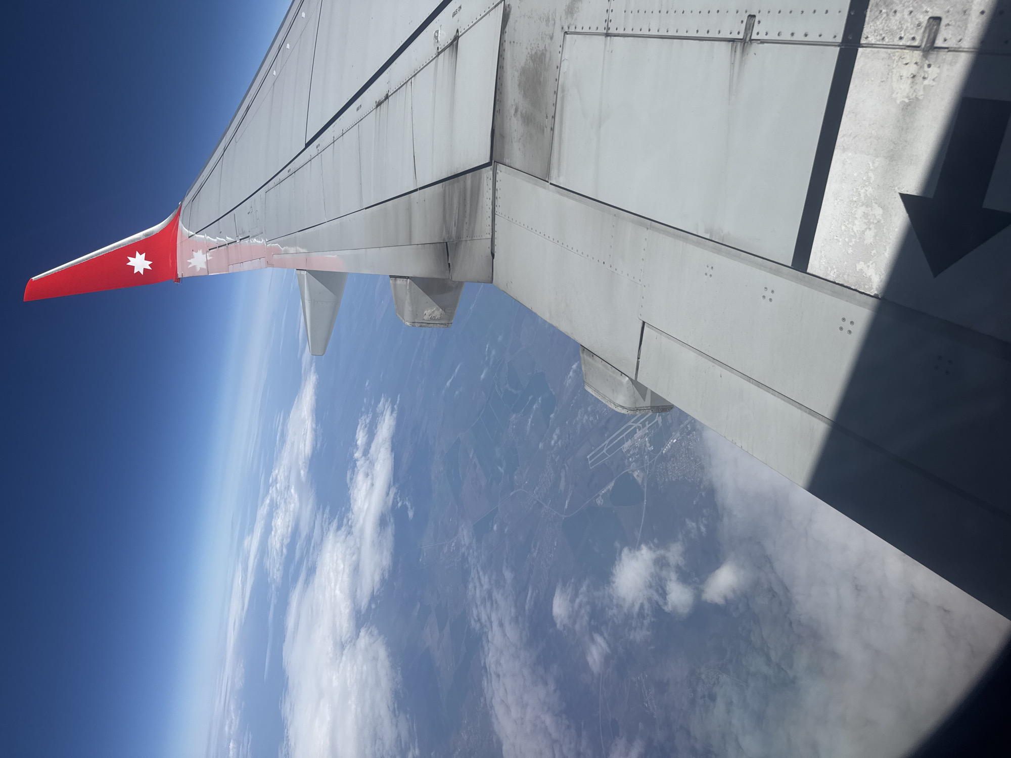 The image shows an airplane wing, with a red wingtip displaying a white star, extending over a landscape. The sky is visible in the background, transitioning from deep blue to a lighter blue closer to the horizon. Below the wing, there are visible land features including fields, possible settlements, and a network of roads. Cloud formations are also visible over the landscape. The angle of the shot suggests it was taken from inside the aircraft. There is no information confirming if the plane is flying near or over the city of Забегалово, Russia.
