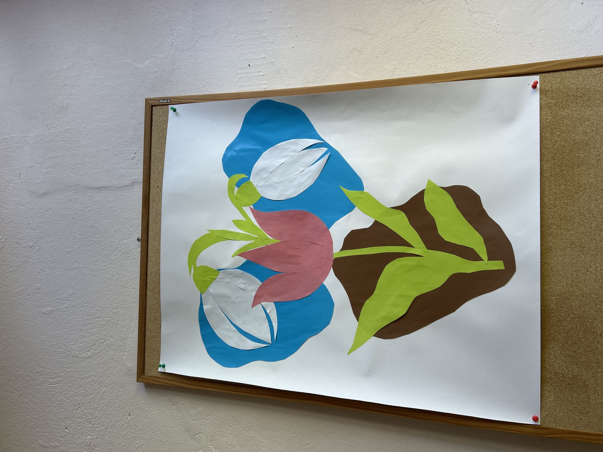 The image shows a corkboard with a framed white sheet of paper pinned to it. The paper features a collage depicting stylized tulips. The collage is composed of cut paper shapes in various colors: blue (background), white (tulip petals), pink (tulip petals), green (stems and leaves), and brown (background). The scene is located indoors, with a textured, light-colored wall visible in the background. The precise location within Dve Mogili, Bulgaria, cannot be determined from the given image.