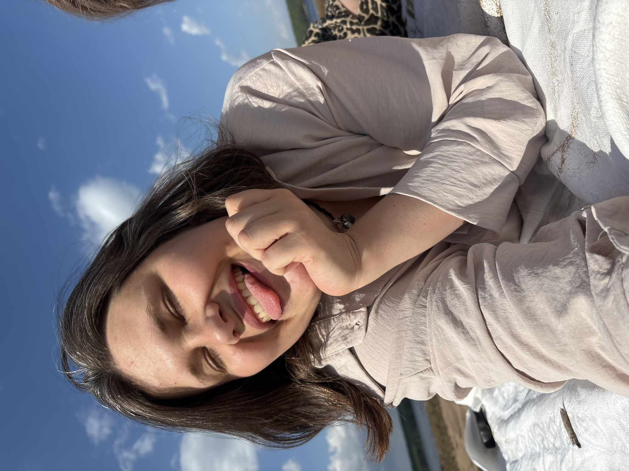 A young woman lies on her side, sticking her tongue out and making a playful gesture with her hand near her face. She has dark hair and is wearing a light-colored shirt. The background shows a blue sky with white clouds, suggesting an outdoor setting. In the distance, there's a glimpse of water and possibly the shore. The setting is likely a park or beach area in Chaikovsky, Russia, based on the surrounding environment.